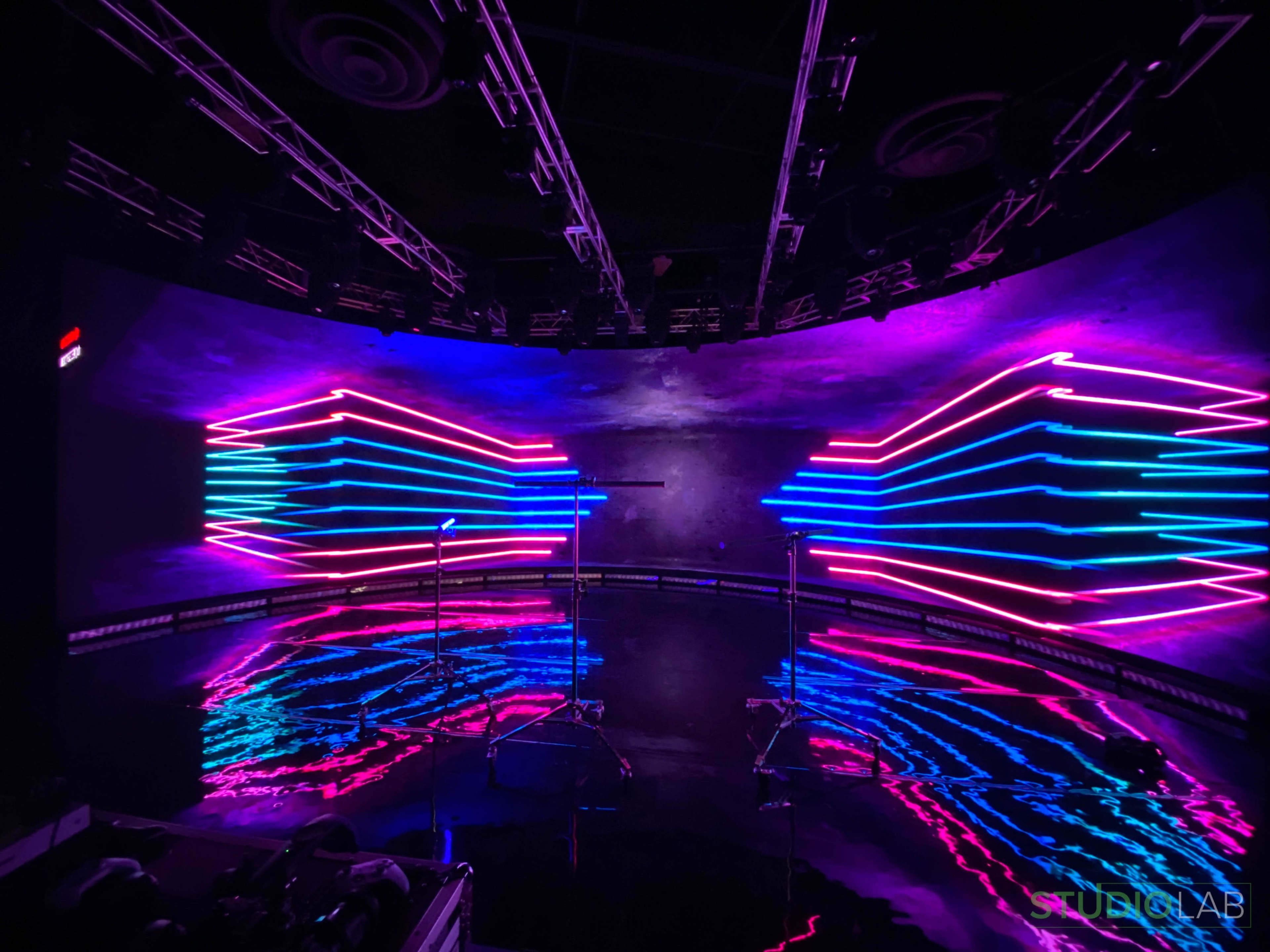 A stage with neon lights arranged in a symmetrical pattern, reflecting off a glossy floor.