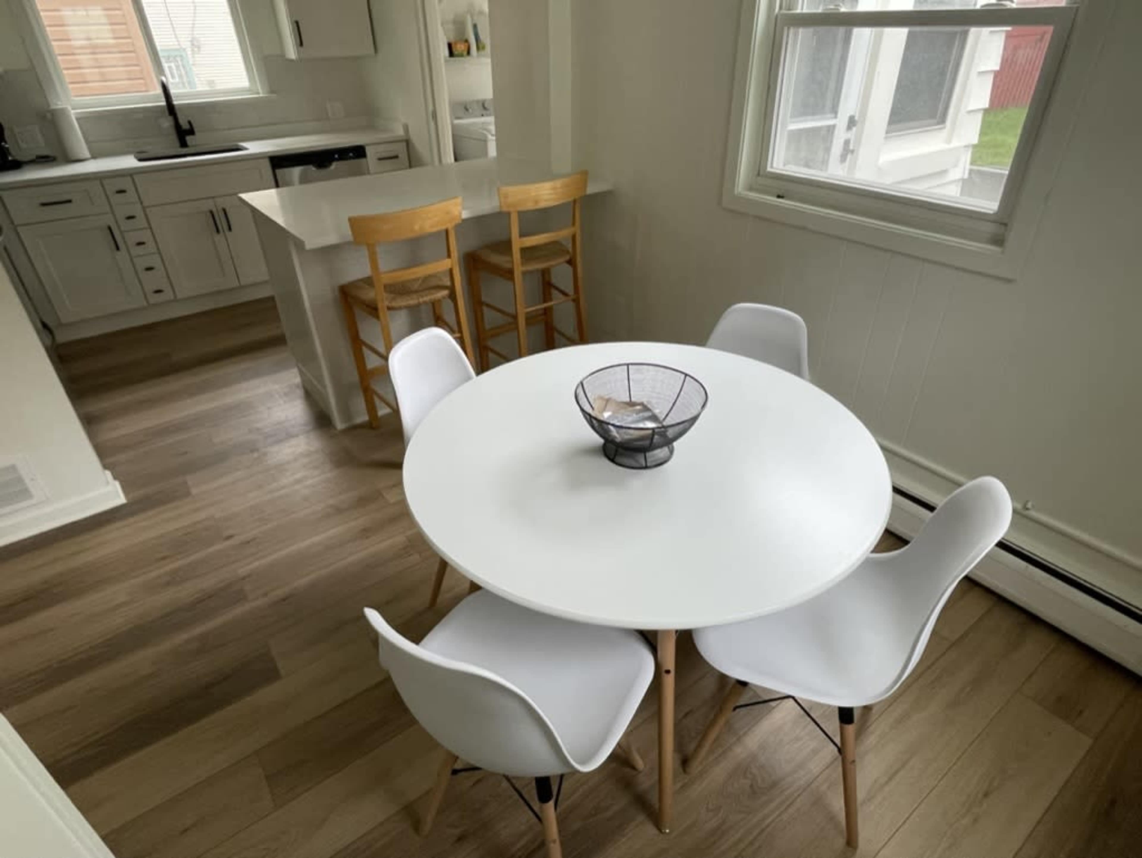 A round white table is set with a bowl in the center, surrounded by four white chairs, in a bright kitchen area with wooden floors.
