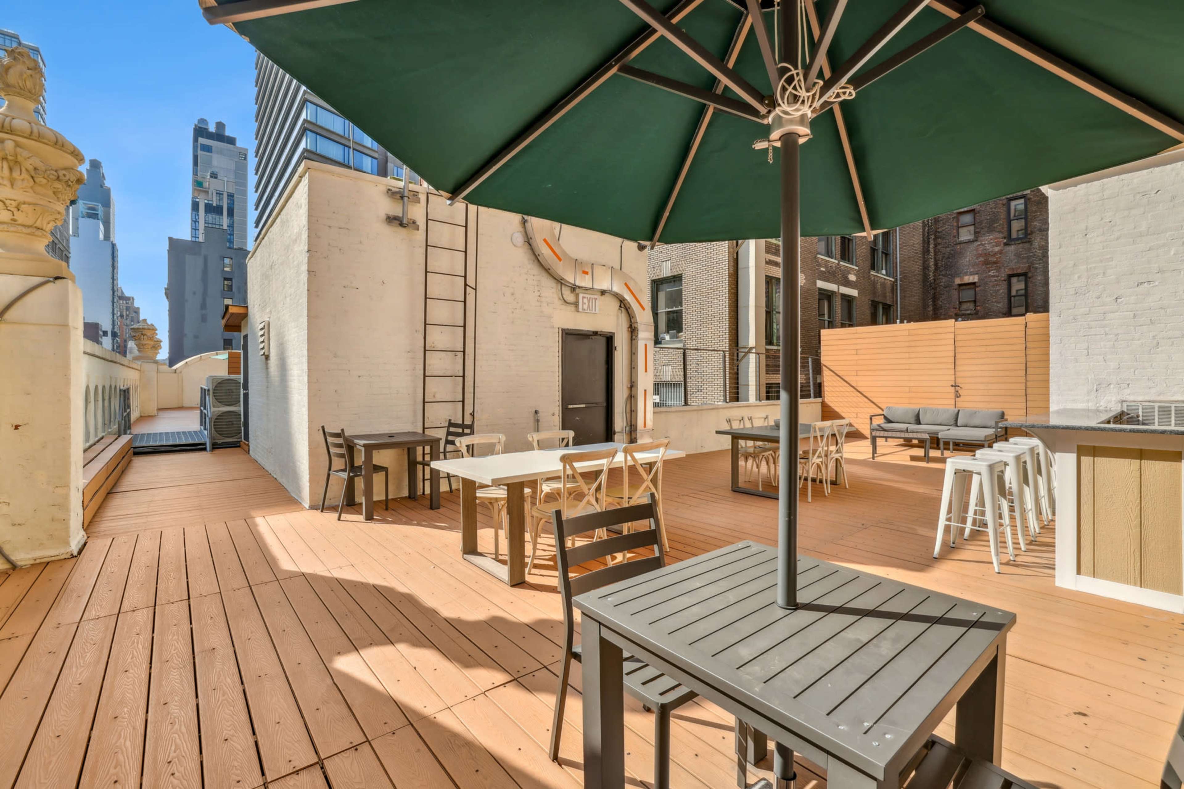 The image shows a rooftop terrace with wooden decking, tables, chairs, and an umbrella, surrounded by buildings in an urban setting.