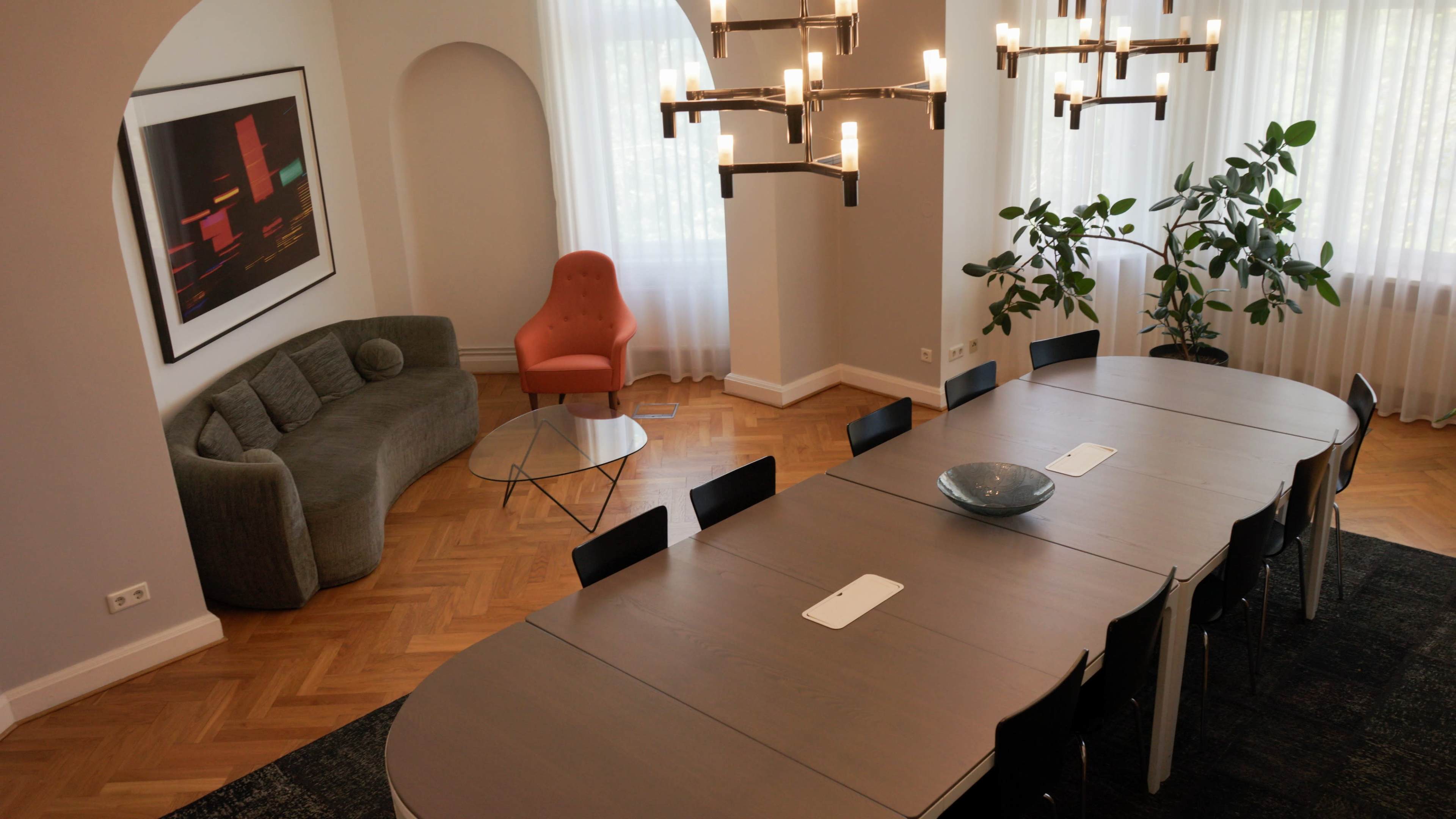 A modern dining area features a large oval table surrounded by black chairs, a gray sectional sofa, an orange accent chair, and a chandelier above, with bright natural light coming from large windows.