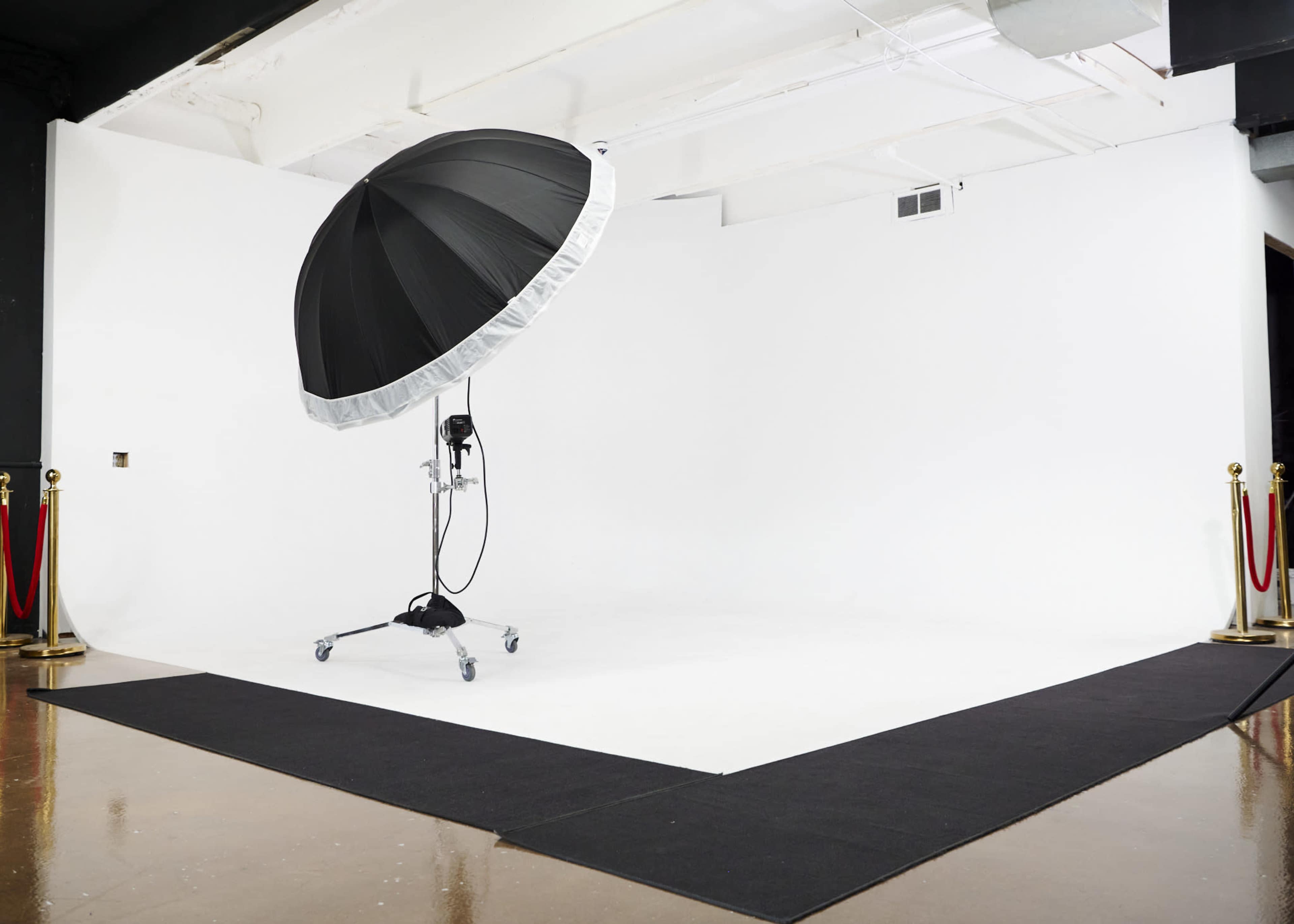 A photography studio features a large black umbrella softbox on a stand, positioned on a white backdrop with a black mat in front.