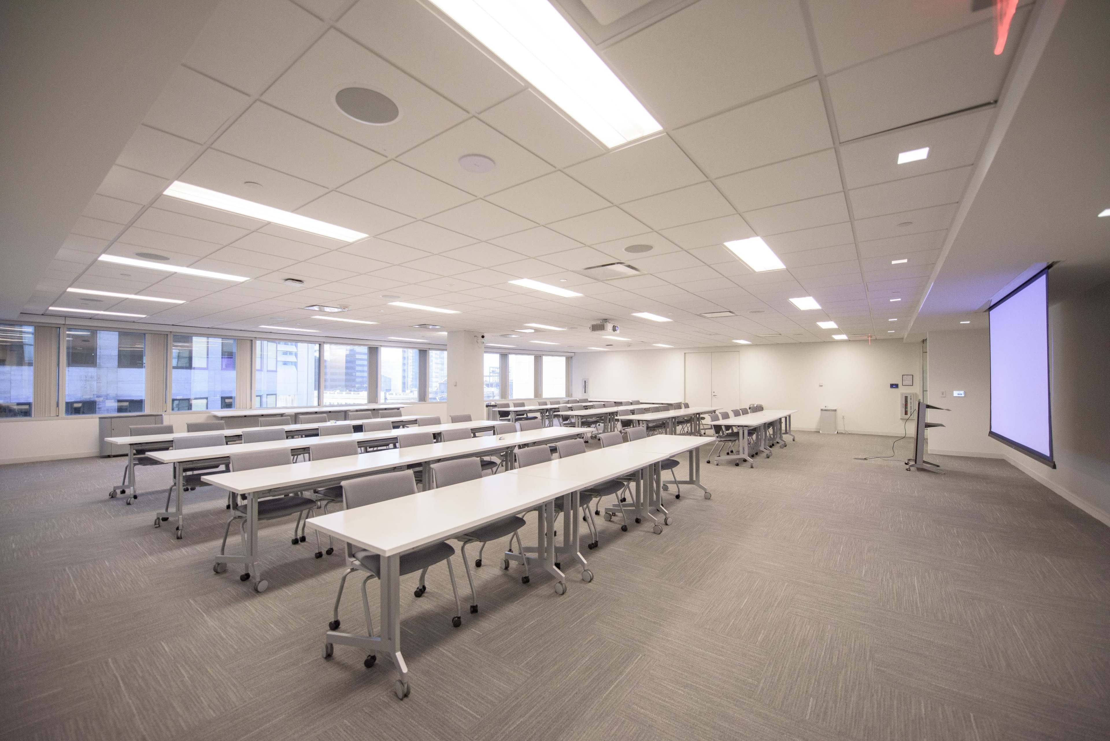 A spacious conference room features multiple tables arranged in rows, with a large screen at one end and windows providing natural light.