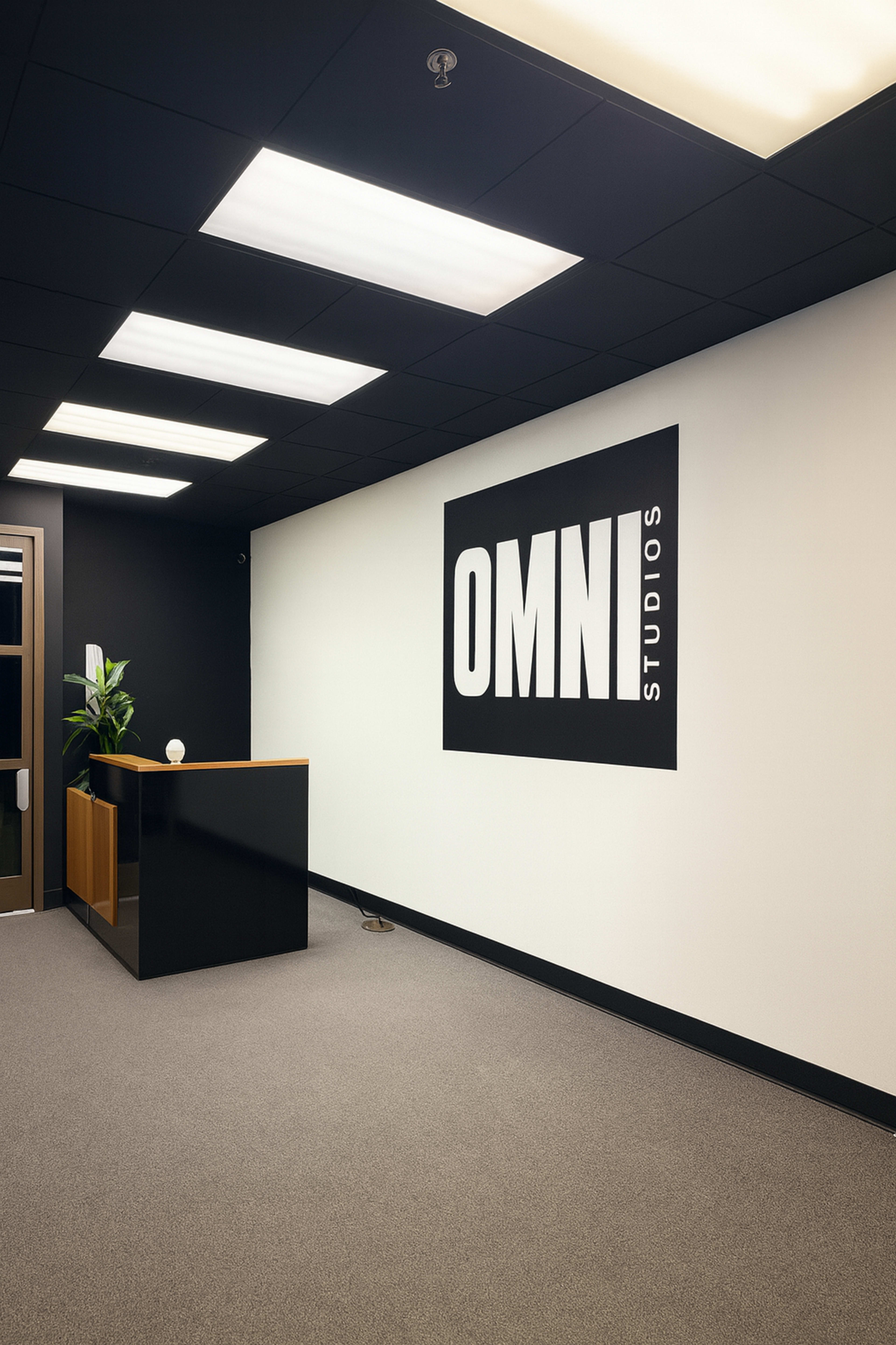 The image shows a modern office reception area with a dark ceiling, a light carpet, a wooden reception desk, and a large black and white sign on the wall that reads "OMNI Studios."