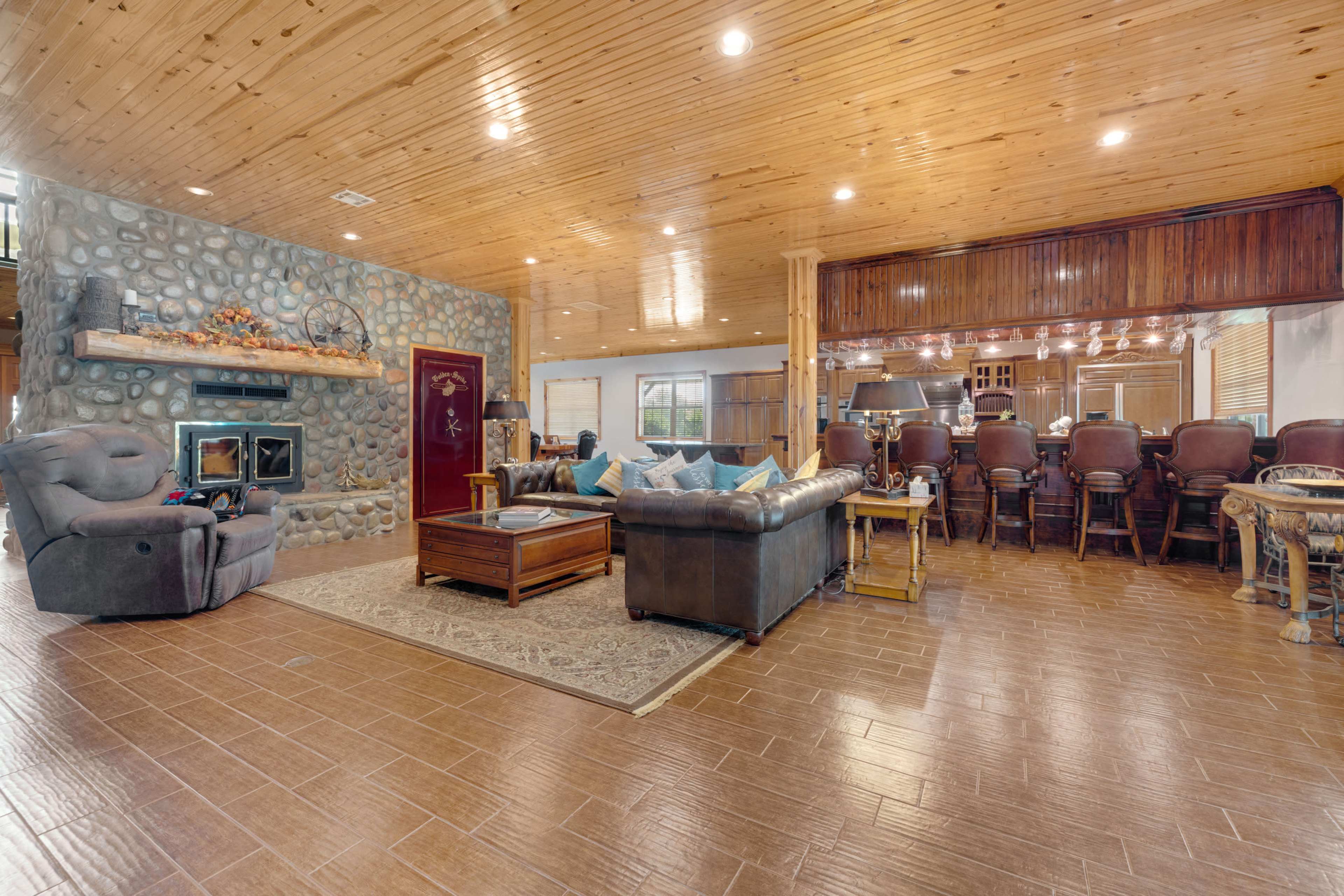 A spacious living room with a stone fireplace, wooden ceiling, and a seating area arranged around a coffee table, adjacent to a kitchen with a bar.