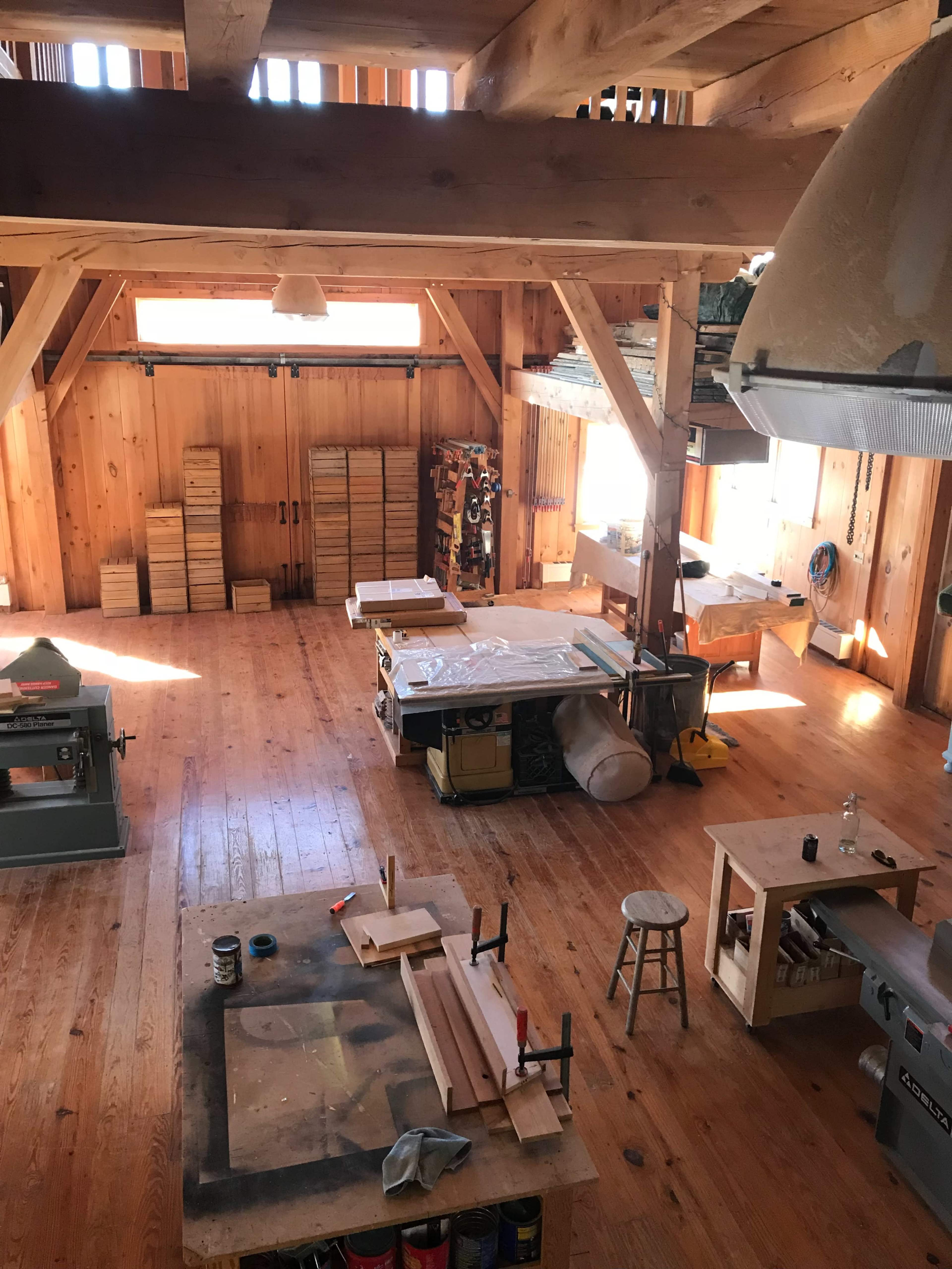 The image shows a spacious woodworking shop with wooden beams, tools, and workstations arranged neatly in a sunlit environment.