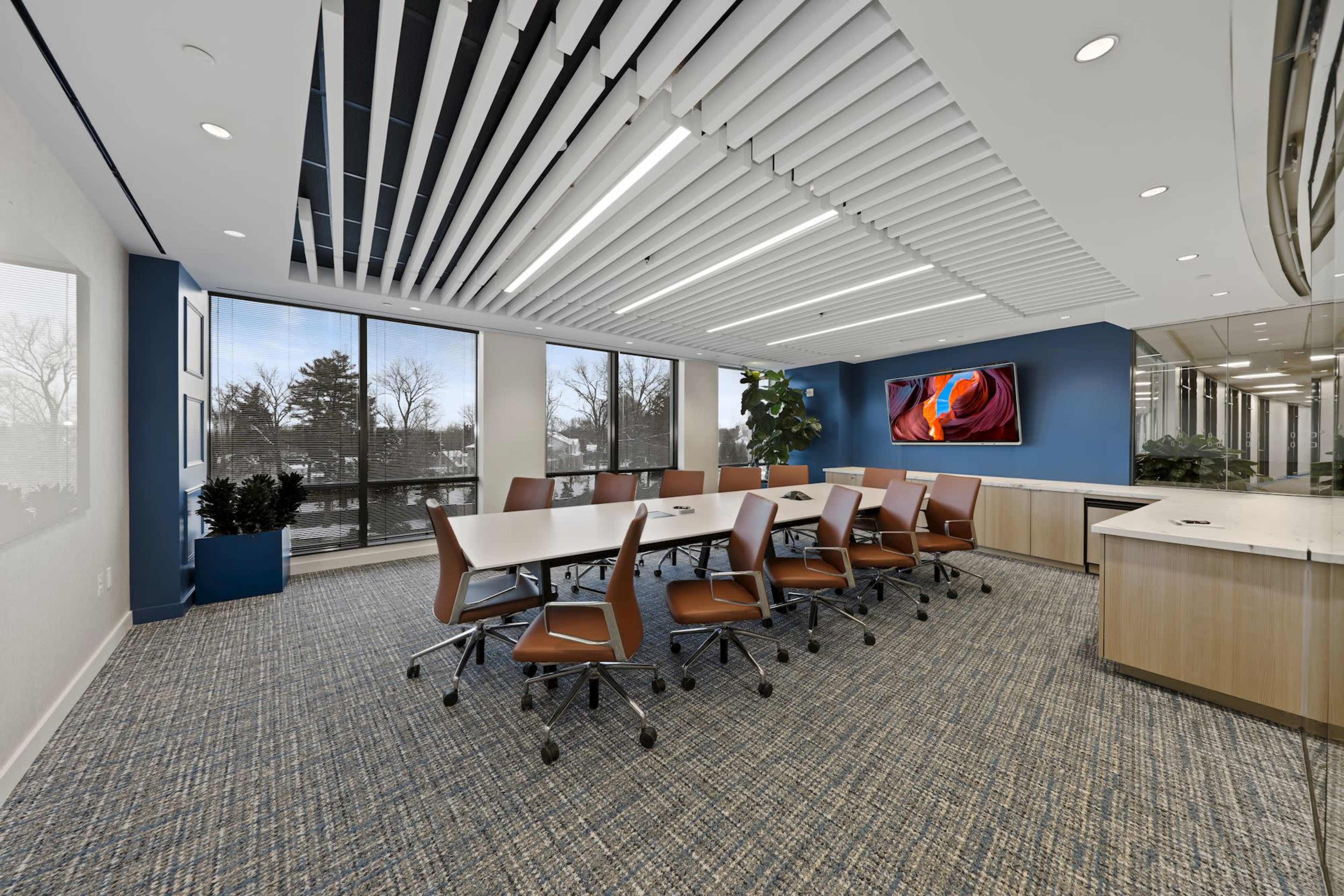 A modern conference room features a large table surrounded by ergonomic chairs, with windows providing natural light and a wall-mounted screen displaying graphics.