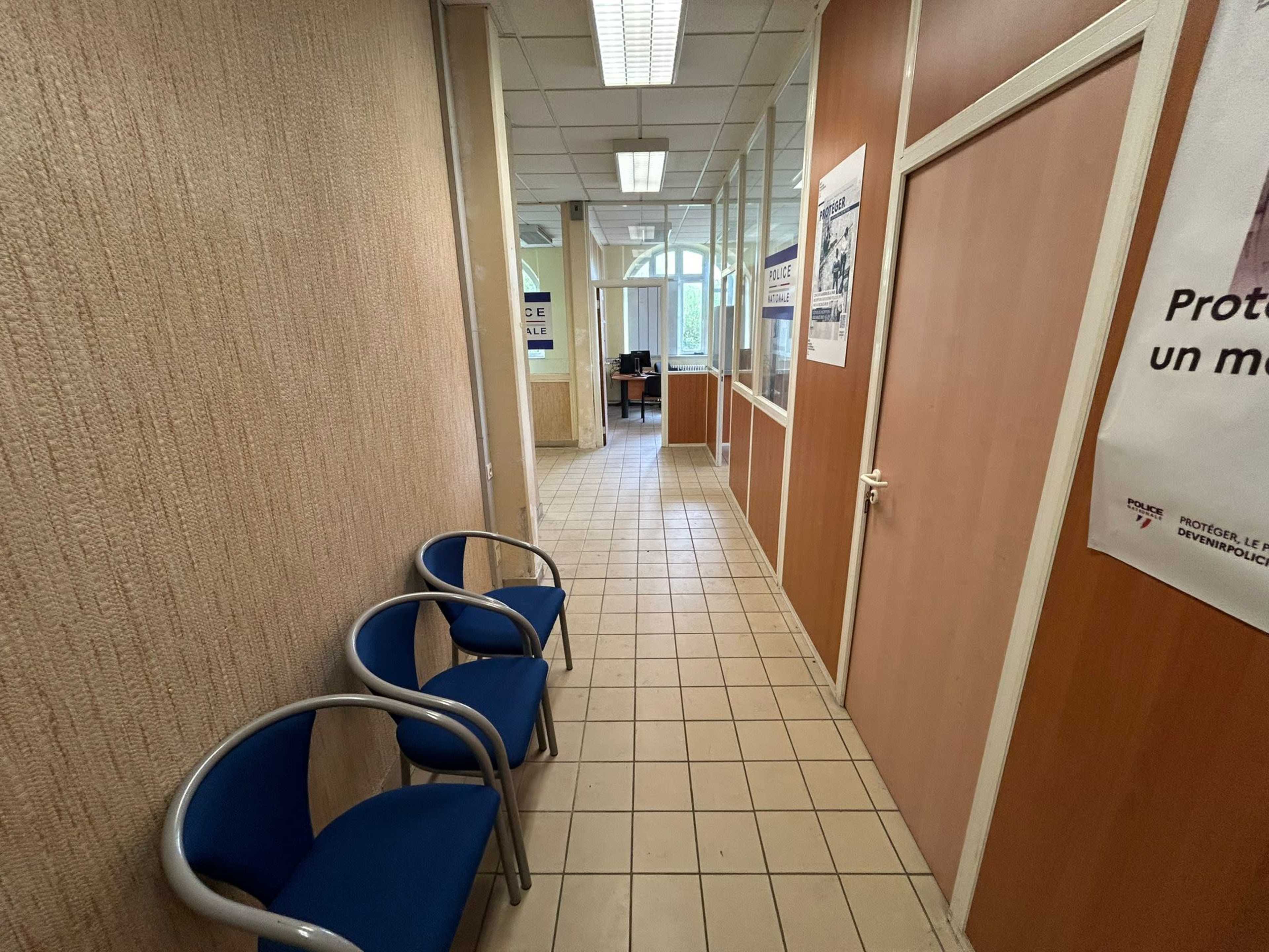 A narrow hallway in an office building, featuring two blue chairs on one side and glass partitions along the length of the corridor.