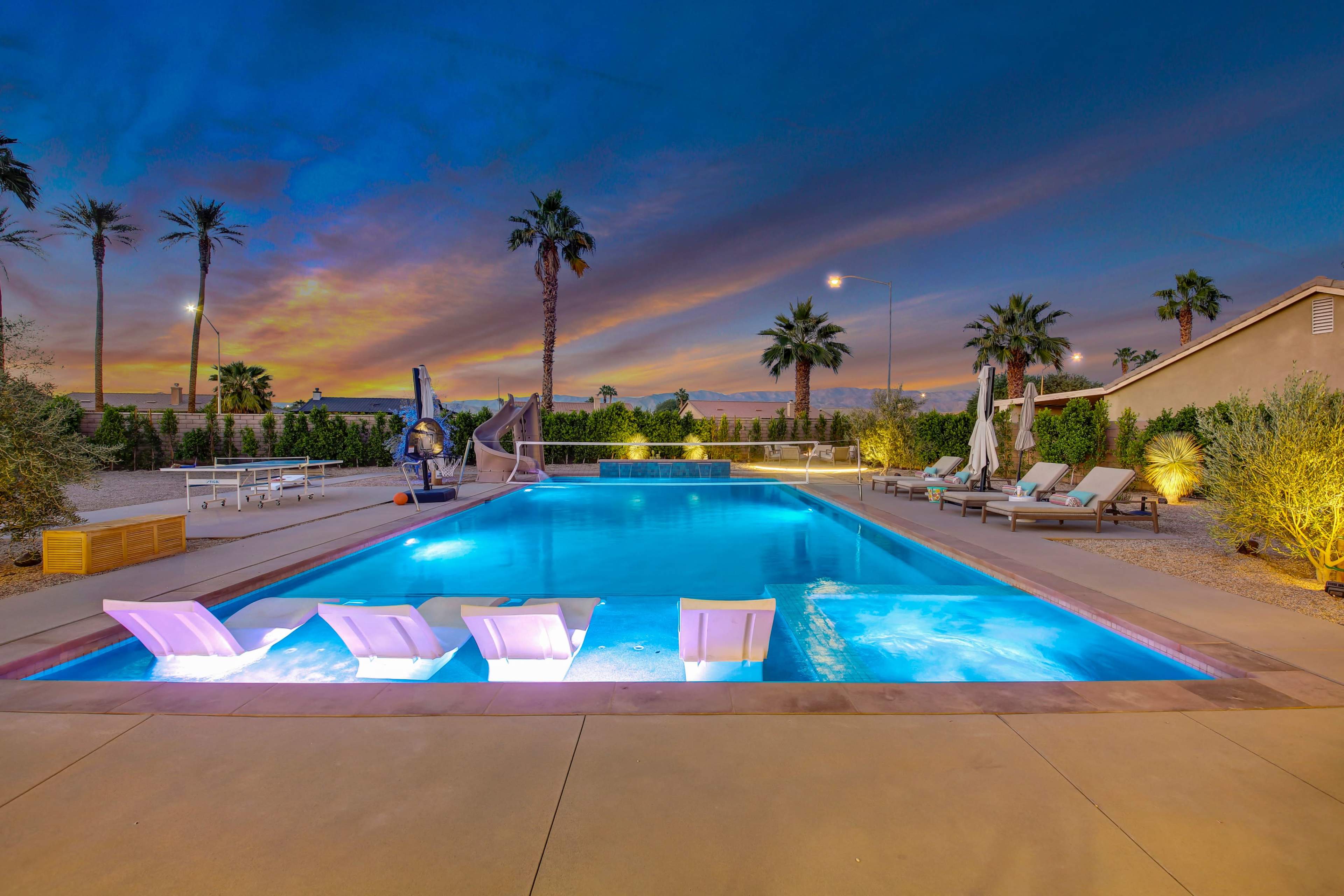 A landscaped pool area featuring a large swimming pool with illuminated loungers, surrounded by palm trees and patio furniture against a sunset backdrop.