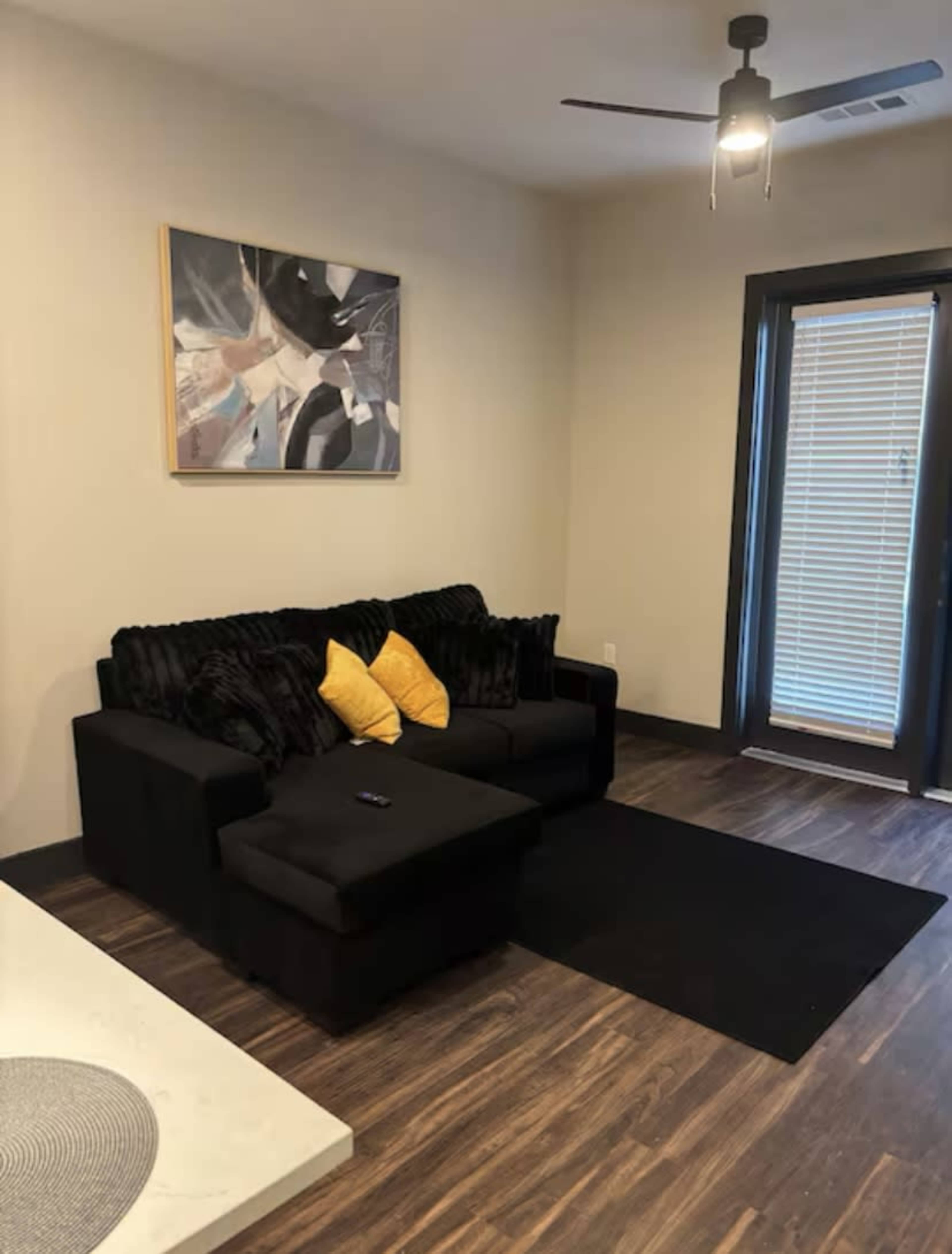 A modern living room features a black couch with yellow pillows, a black rug, and a painting on the wall, complemented by large windows with blinds.