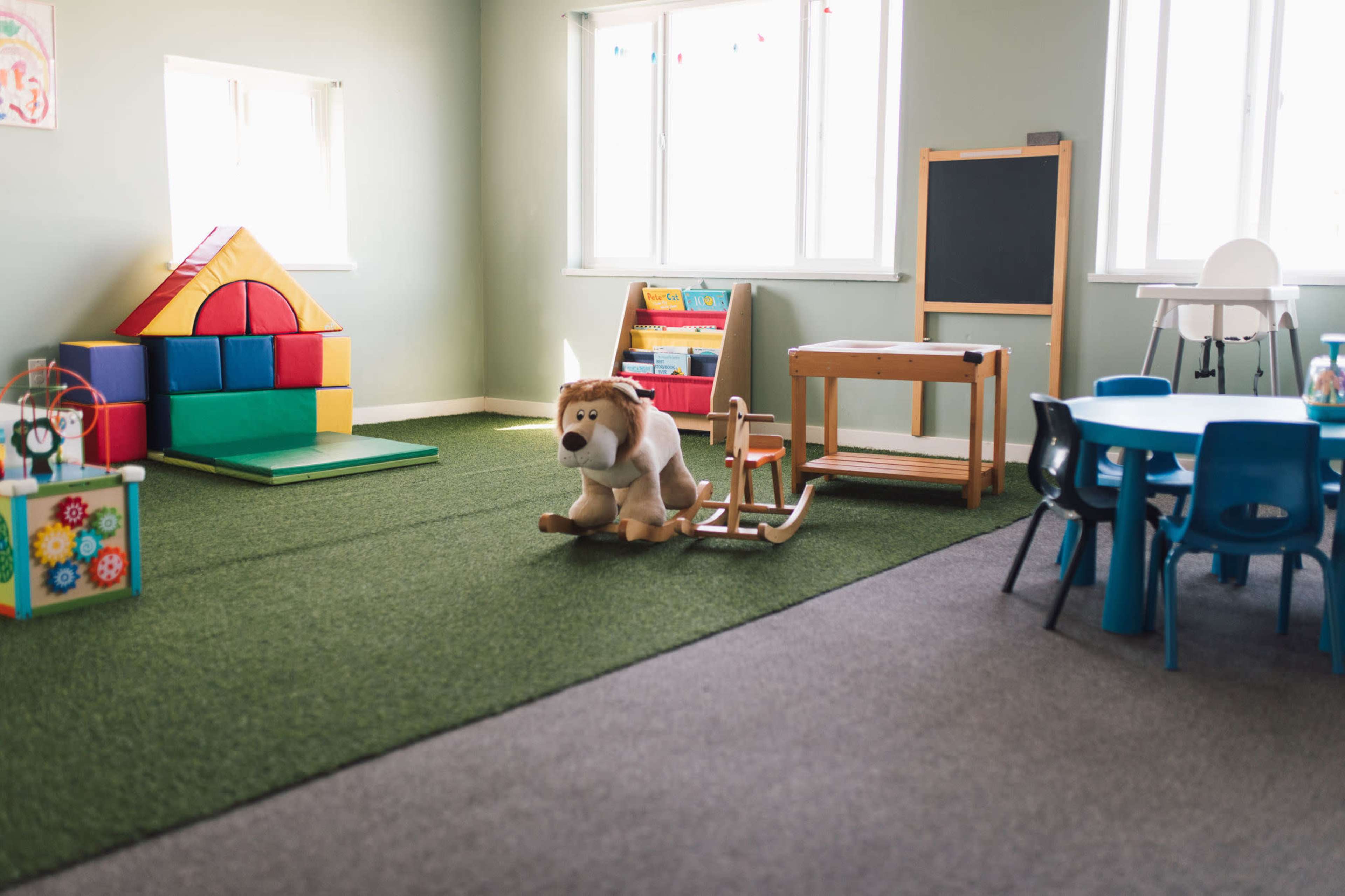 A playroom features colorful building blocks, a plush toy on a rocking horse, a small table with chairs, and a chalkboard.