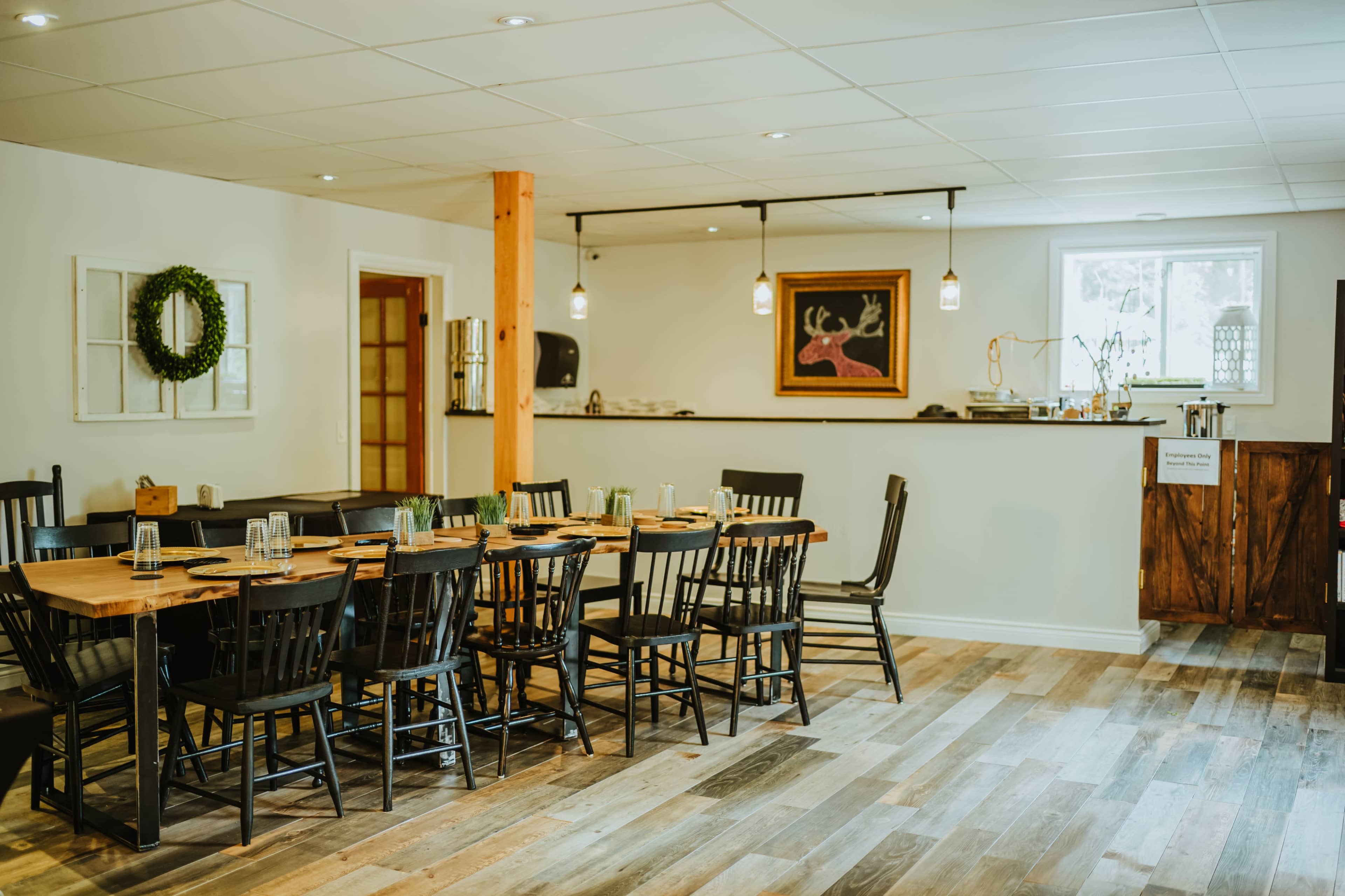 The image shows a simple dining area with a wooden table surrounded by black chairs, a bar counter in the background, and warm, overhead lighting.