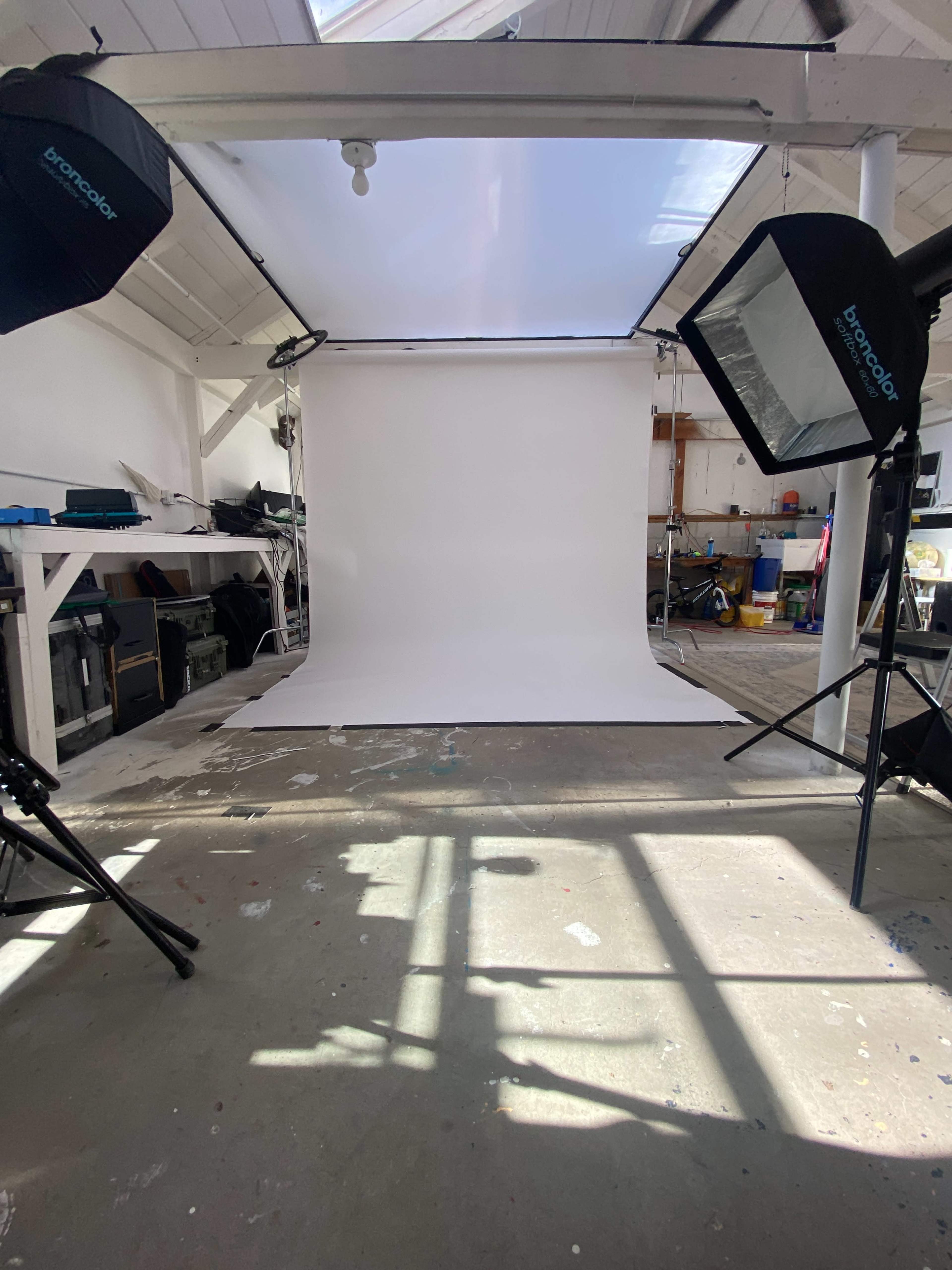 The image shows a photography studio with a white backdrop, two light sources, and various equipment in the background.
