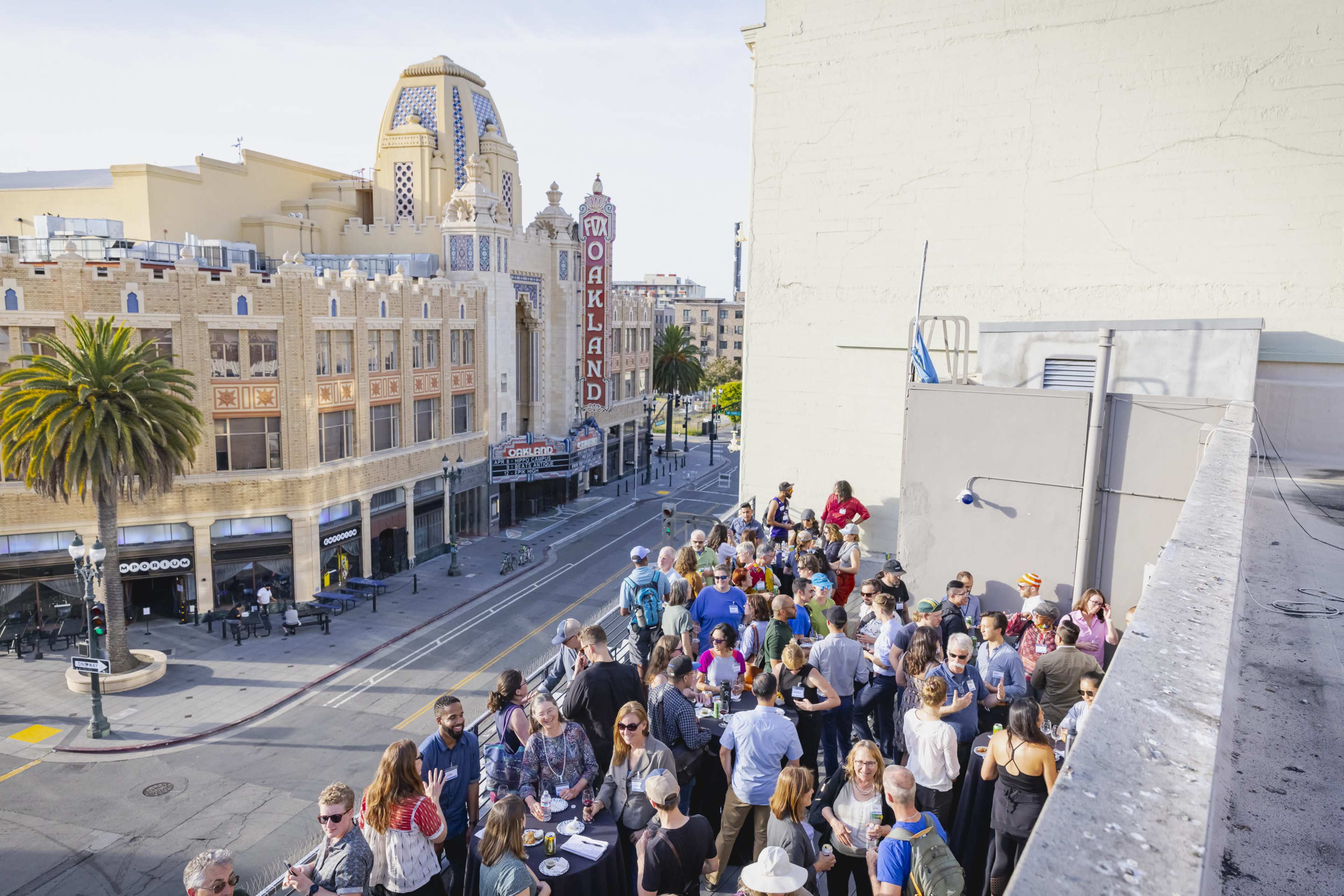 A large group of people gathers on a rooftop overlooking a street in Oakland, with an art deco theater visible in the background.