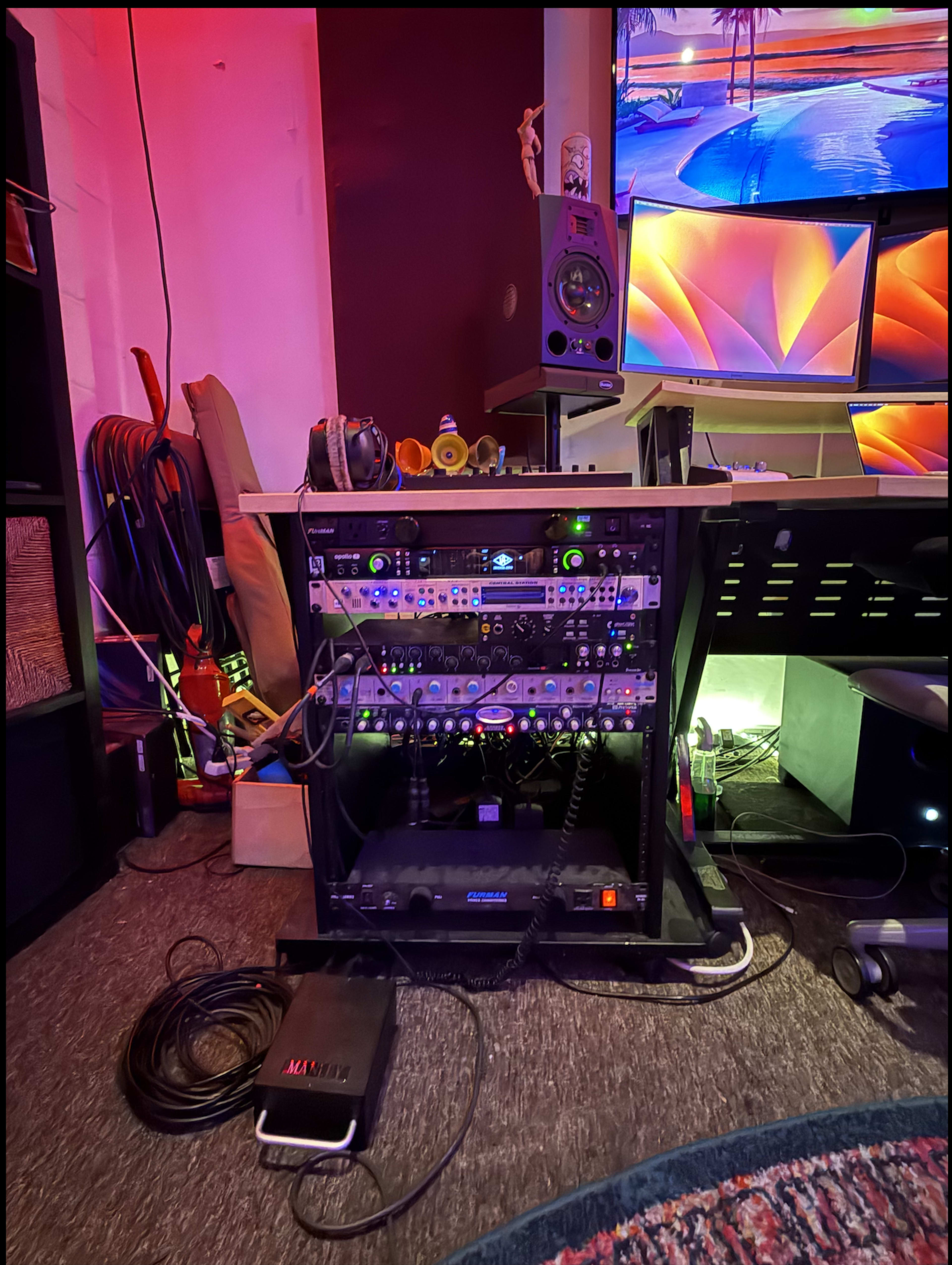 The image shows a well-organized rack of audio equipment with various devices and cables, positioned near a colorful computer monitor setup in a studio environment.