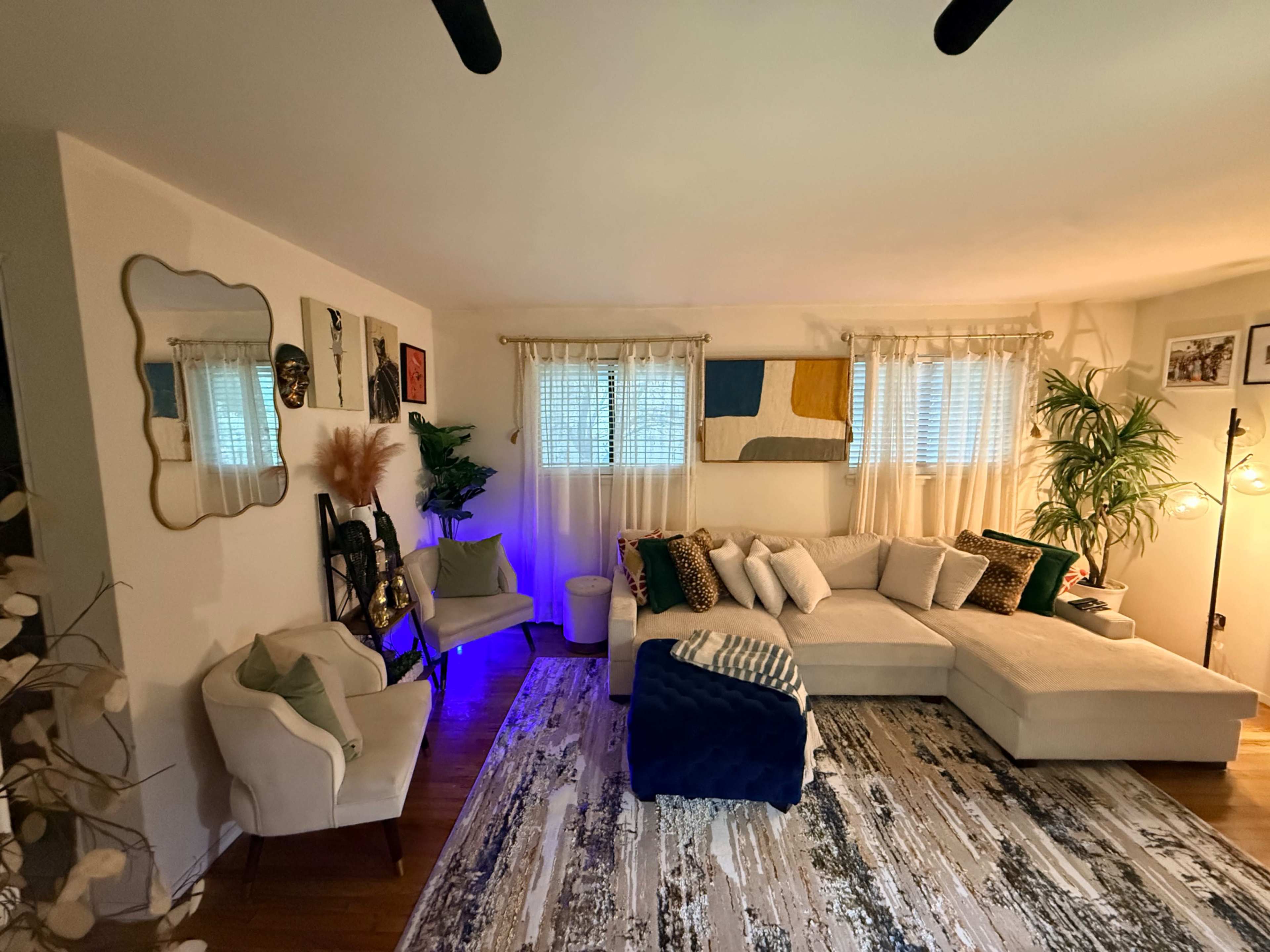 The image shows a cozy living room featuring a large sectional couch, decorative pillows, plants, and a patterned rug, with a warm ambiance created by multiple light sources.