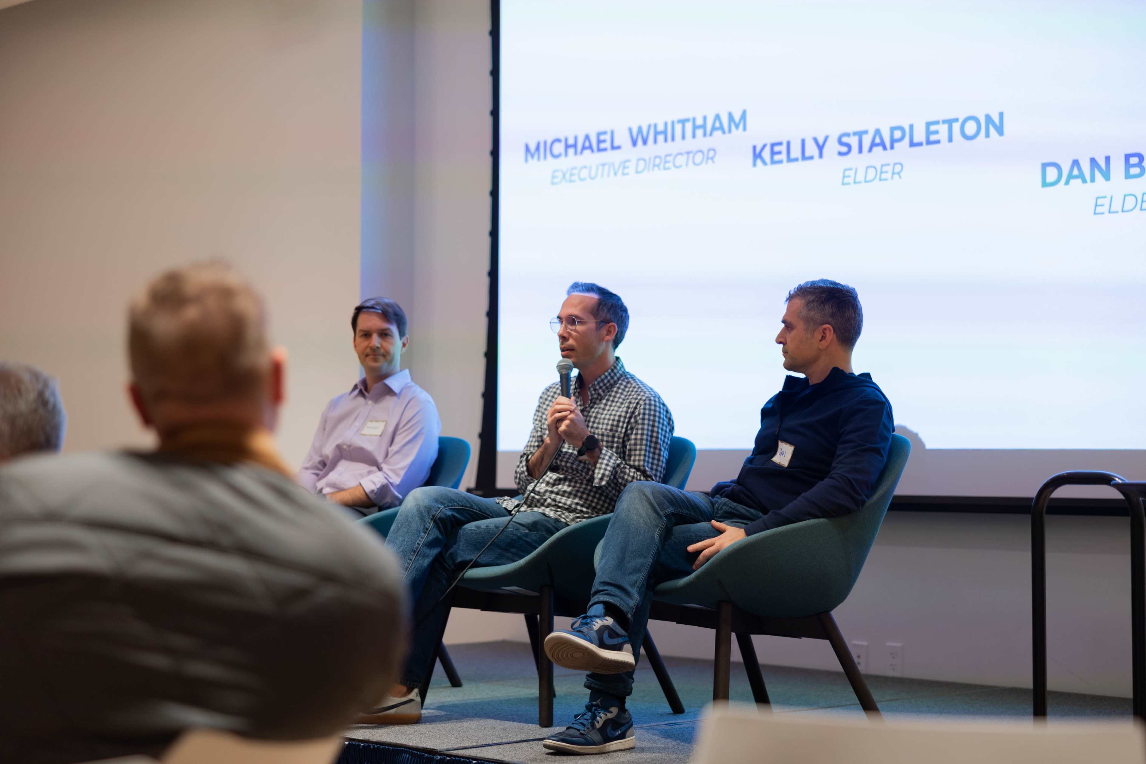 A panel discussion features three speakers seated on chairs, with one person speaking into a microphone while the other two listen.