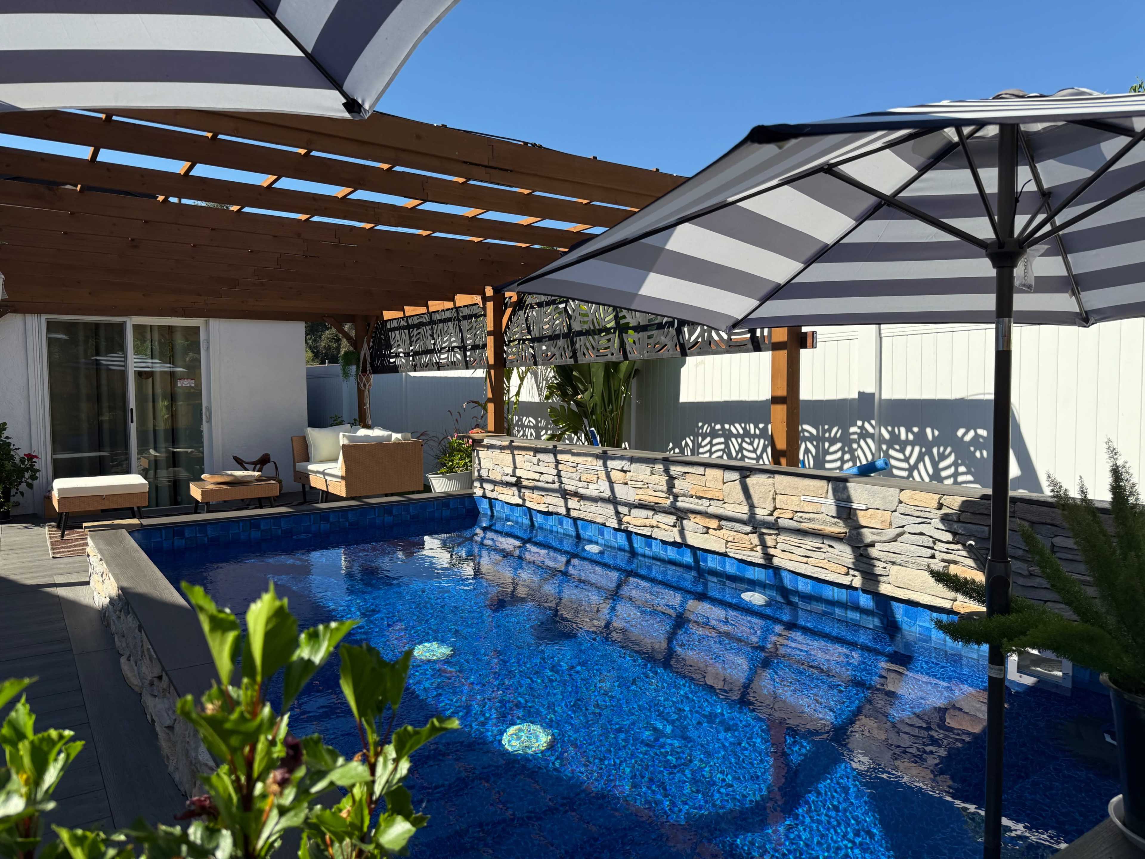 A well-lit backyard features a swimming pool with a stone border, surrounded by lounging chairs and shade umbrellas under a wooden pergola.