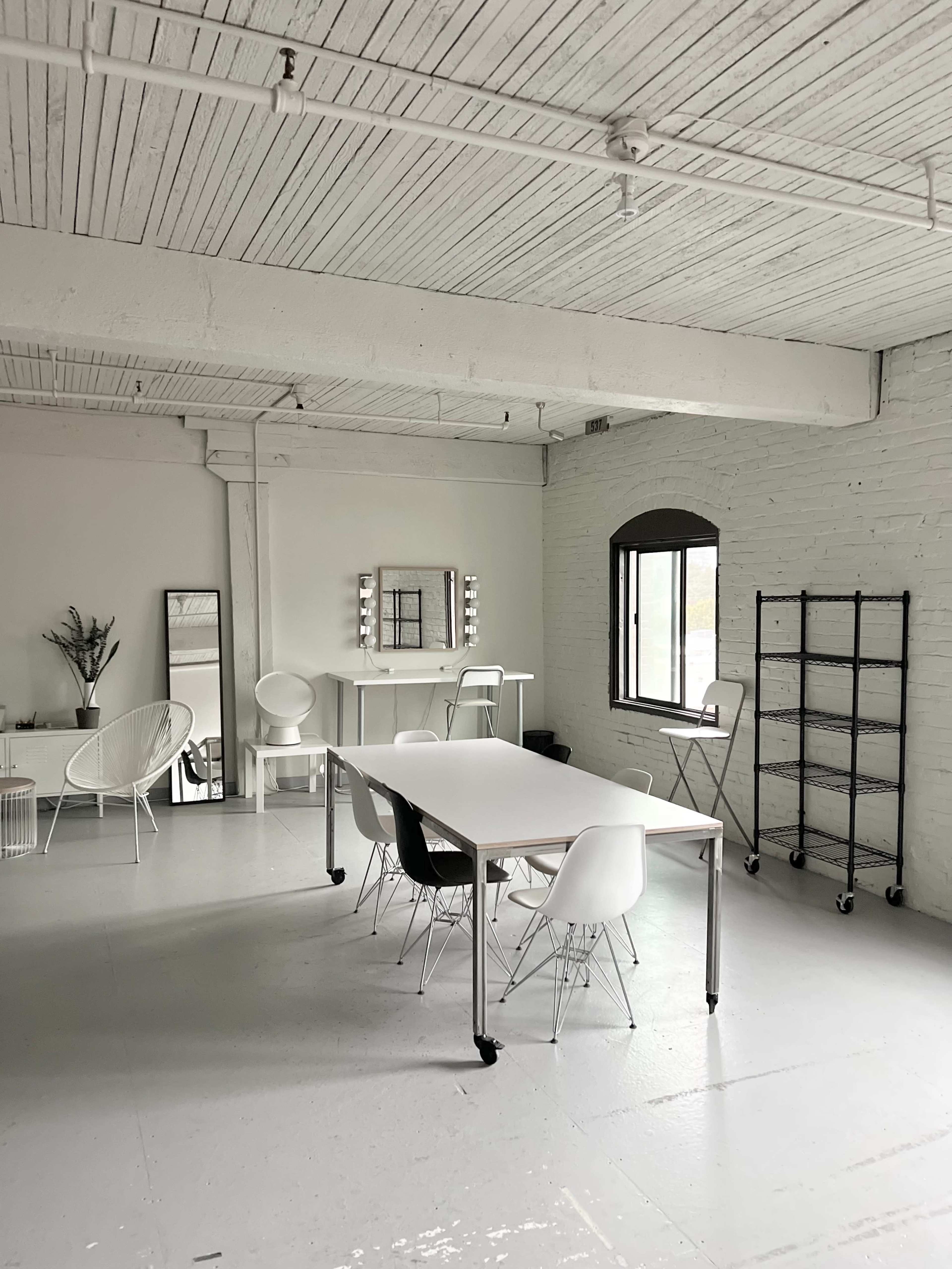 A spacious, well-lit room featuring a long table surrounded by chairs, a mirror on the wall, and a shelving unit.
