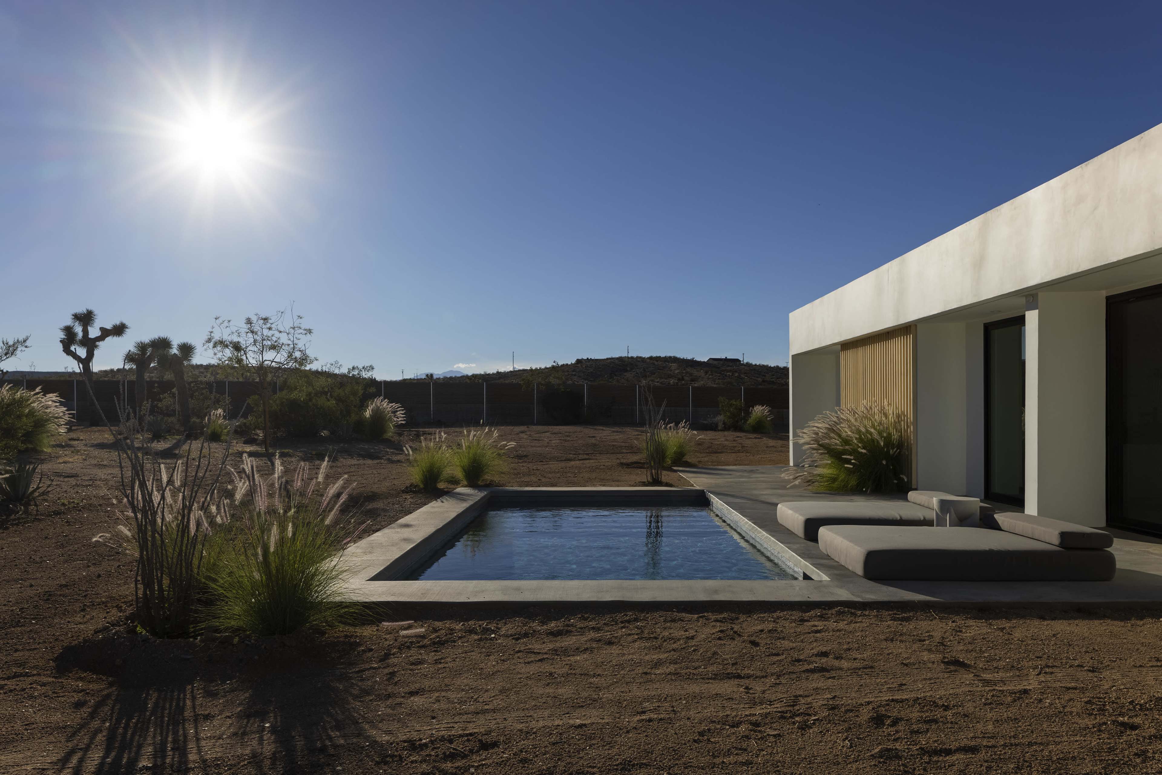 A modern house adjacent to a small, clear swimming pool surrounded by desert landscaping under a bright sun.