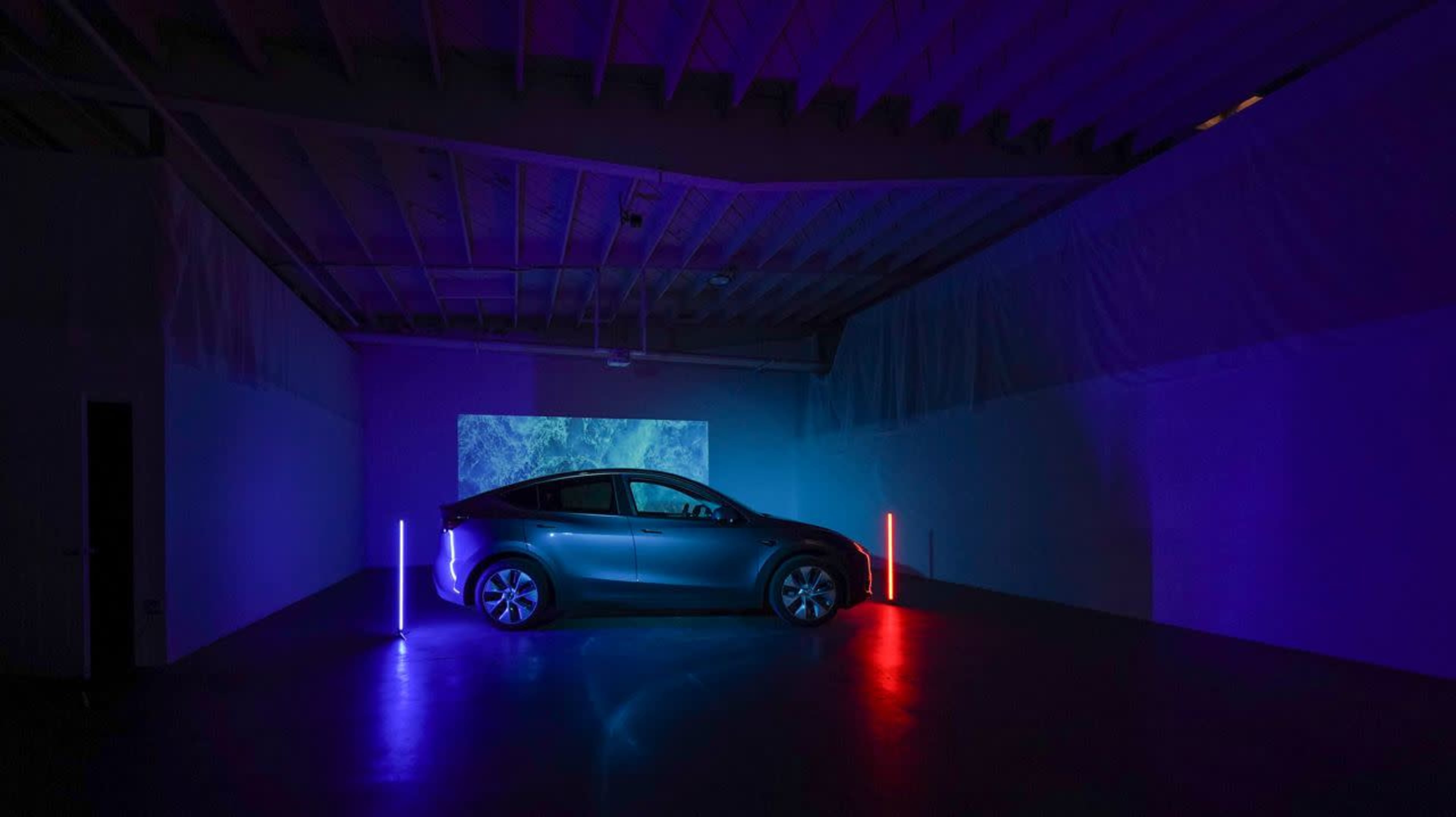 A car is positioned in a dimly lit room, illuminated by purple and blue lights with a projection on the wall behind it.
