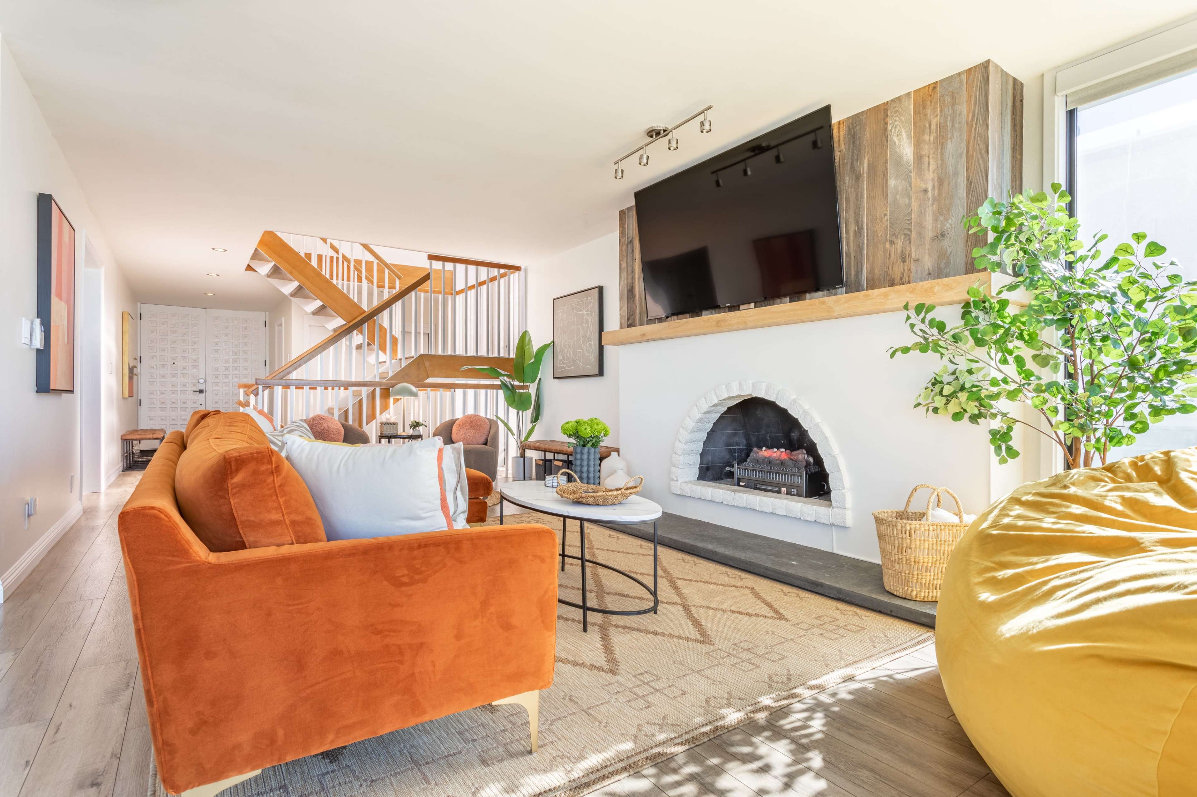 A modern living room featuring an orange couch, a round coffee table, a white fireplace, and a staircase in the background.
