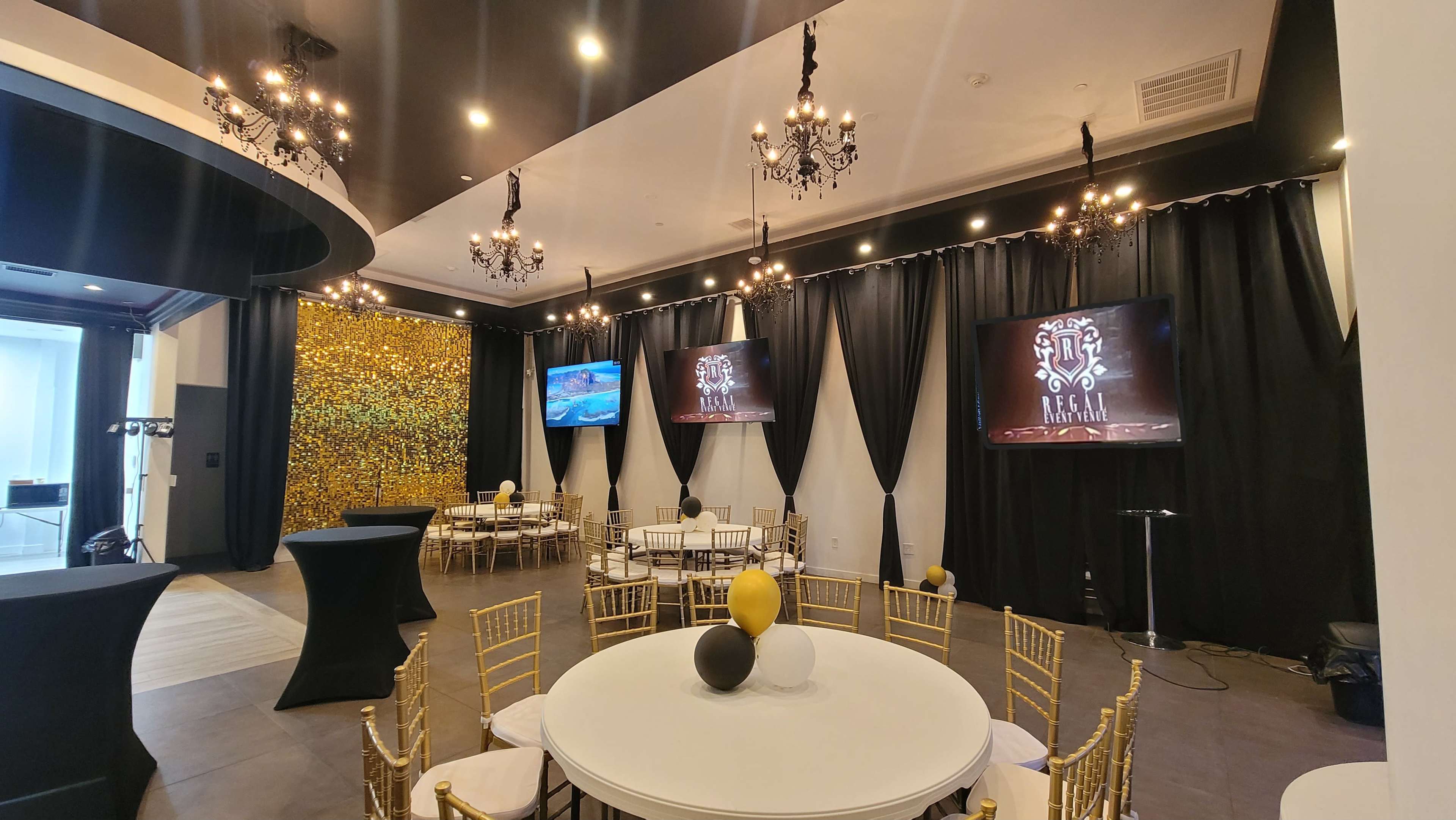 The image shows a spacious event venue with black drapes, golden wall accents, chandeliers, and round tables set with gold and black decorations.