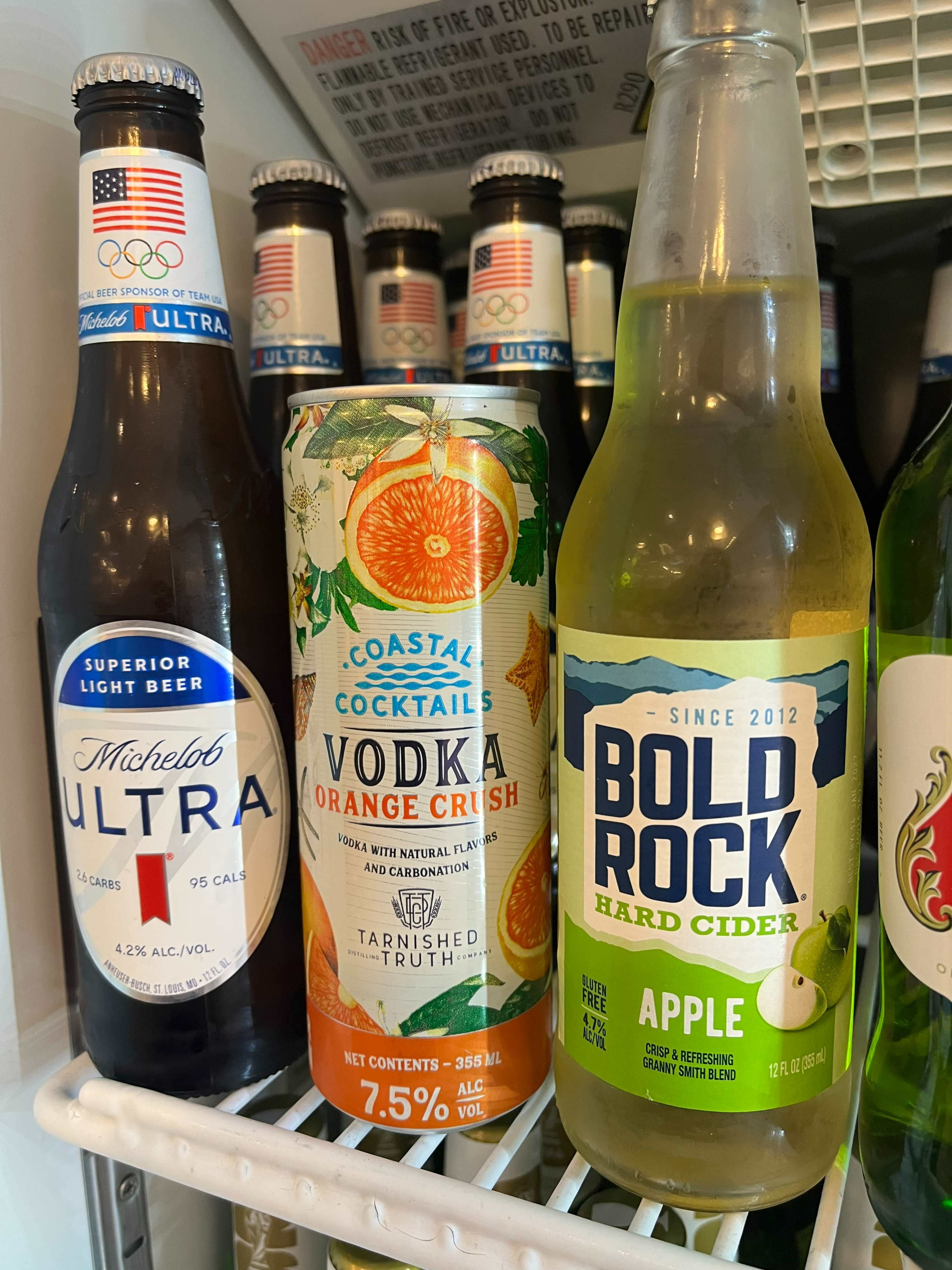 The image shows a refrigerator filled with various alcoholic beverages, including Michelob Ultra beer, a can of Coastal Cocktails Vodka Orange Crush, and a bottle of Bold Rock Hard Cider.