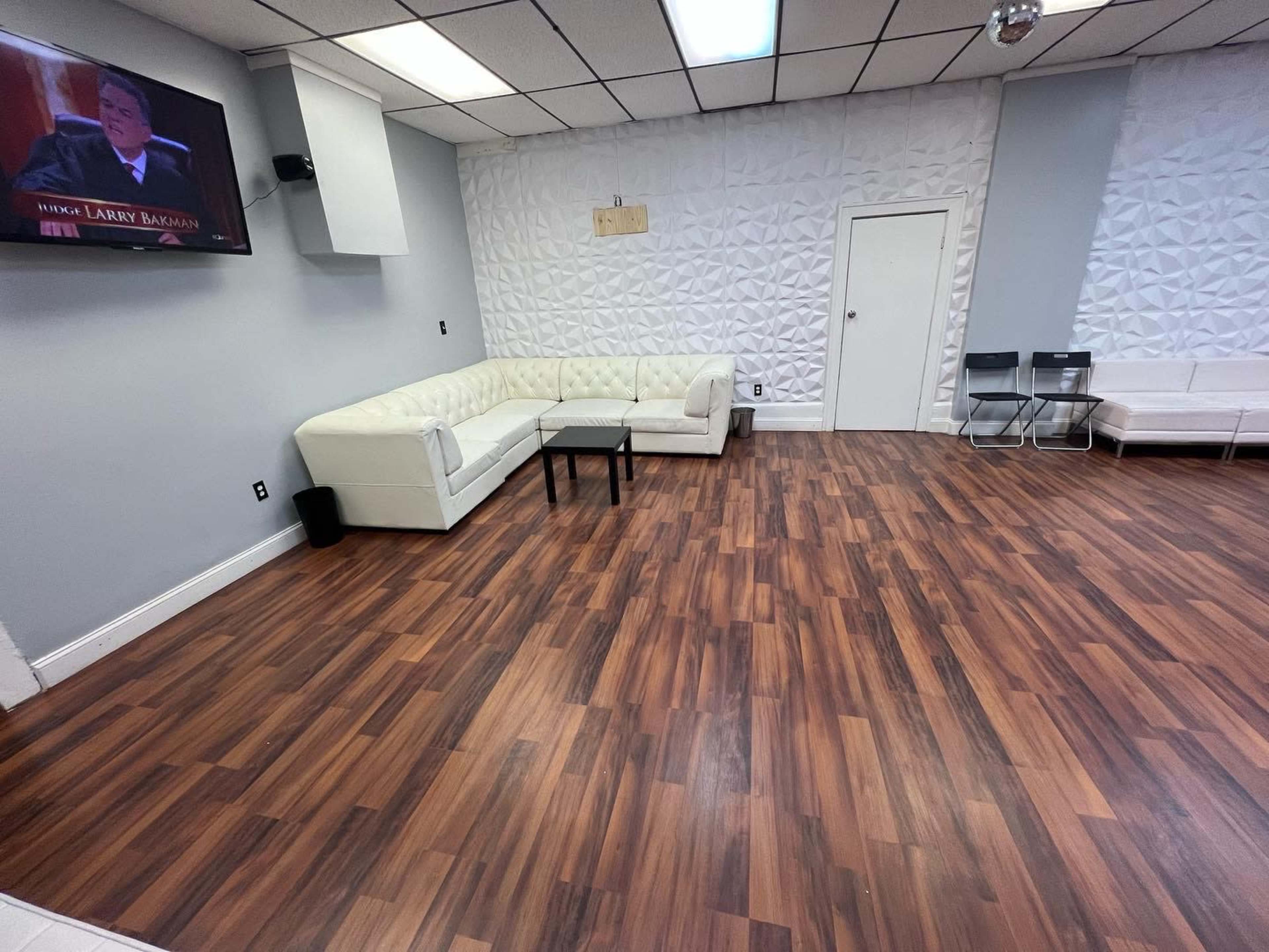 A sparsely furnished room with a white sectional couch, a television mounted on the wall, and a wooden floor.
