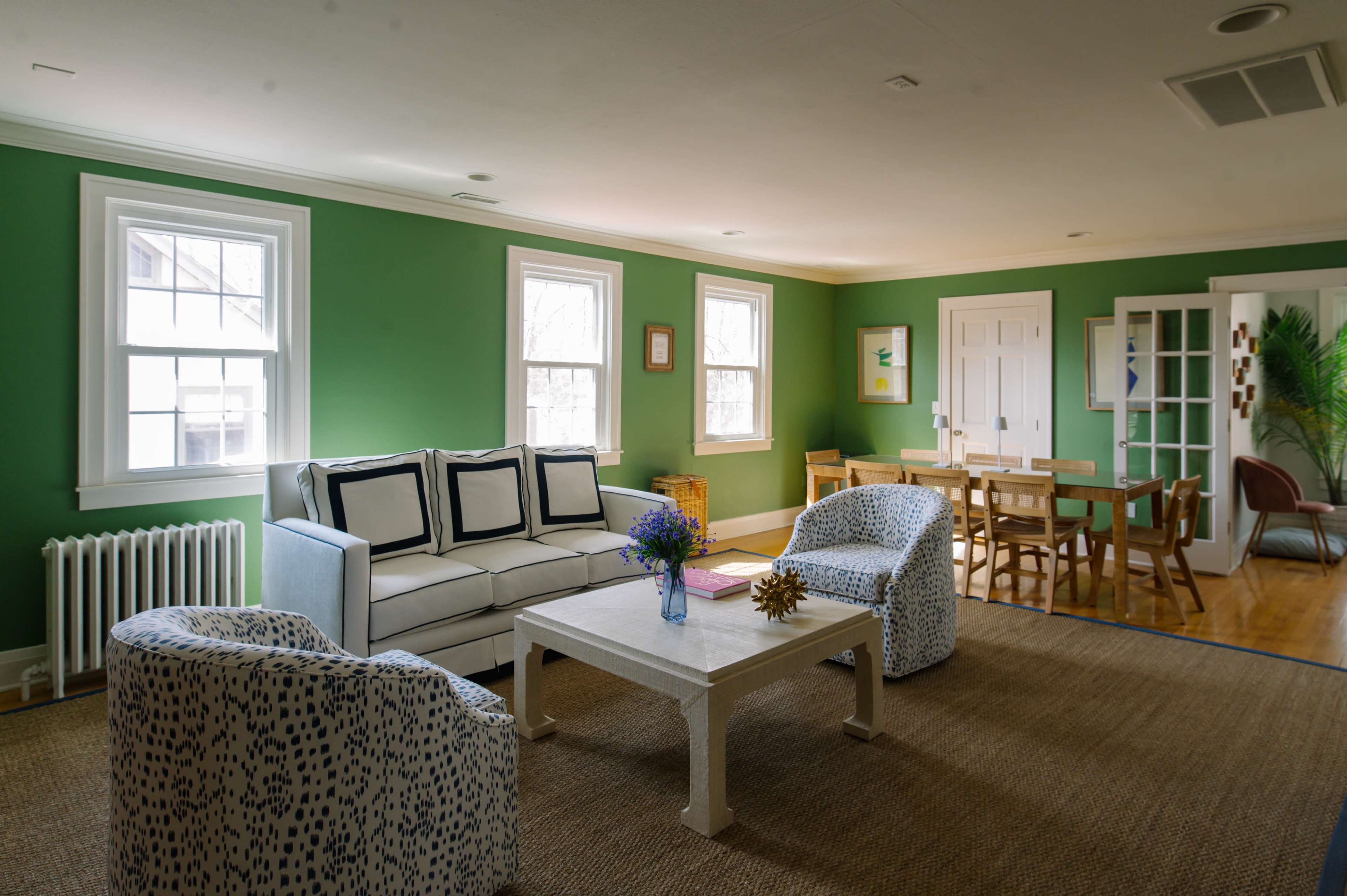 A brightly lit living room with green walls, a white sofa, two patterned chairs, and a table in the center, leading into a dining area with wooden chairs and a door to another room.