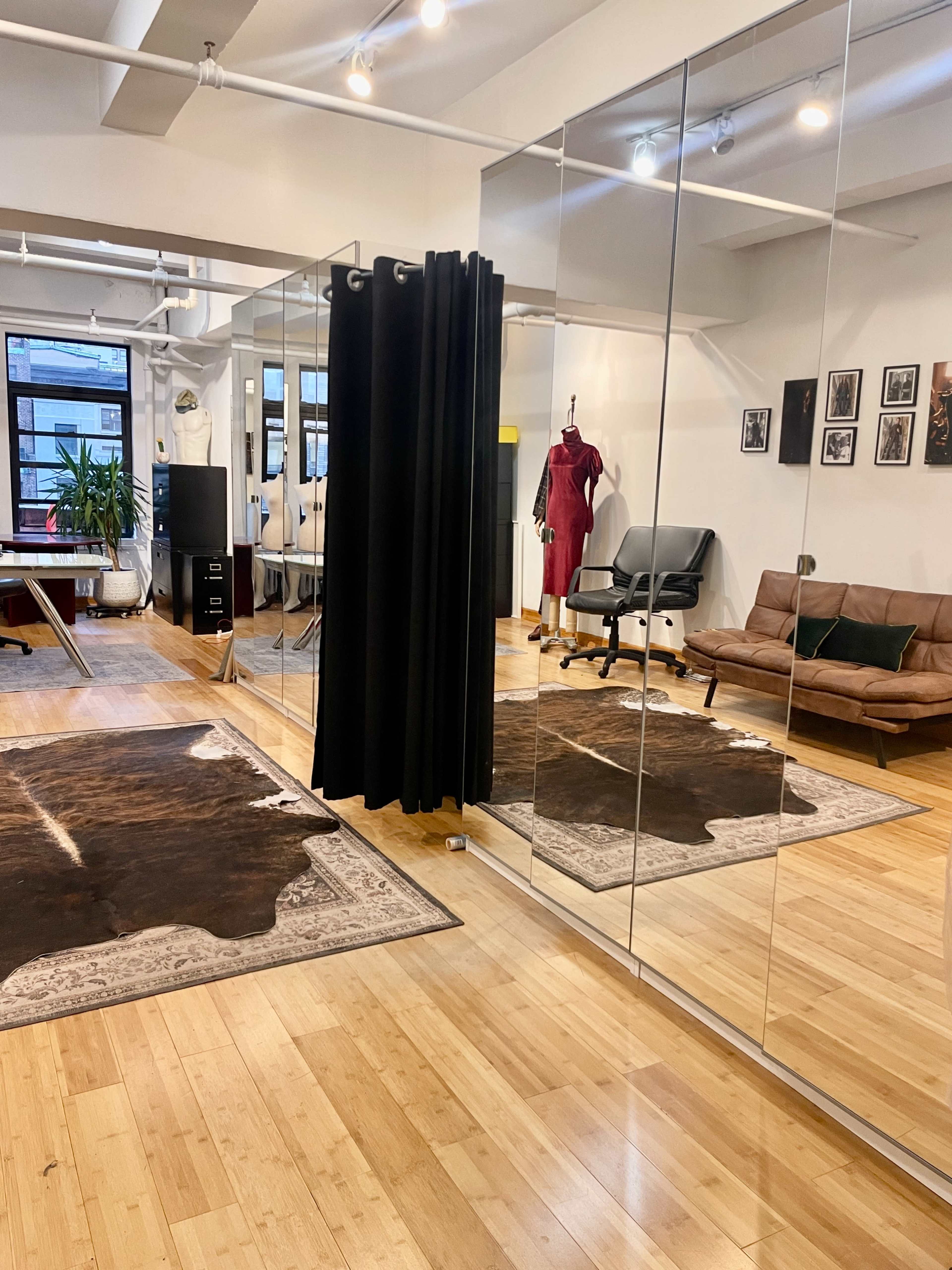 The image shows a spacious studio with mirrors, a clothing rack, a desk chair, a brown sofa, and a large area rug on wooden flooring.