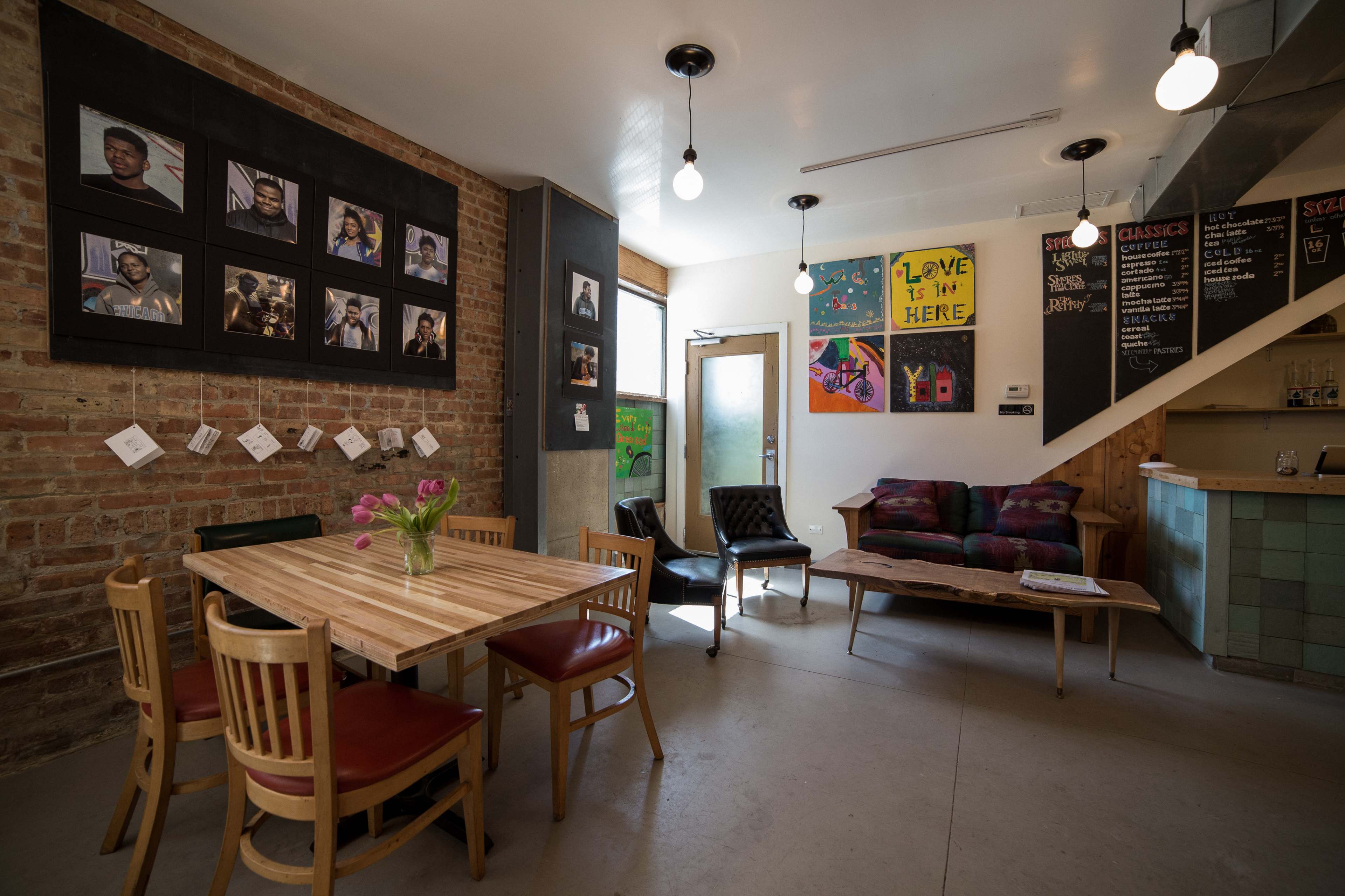 A cozy café featuring a wooden table surrounded by chairs, colorful artwork on the walls, and framed photographs depicting people.