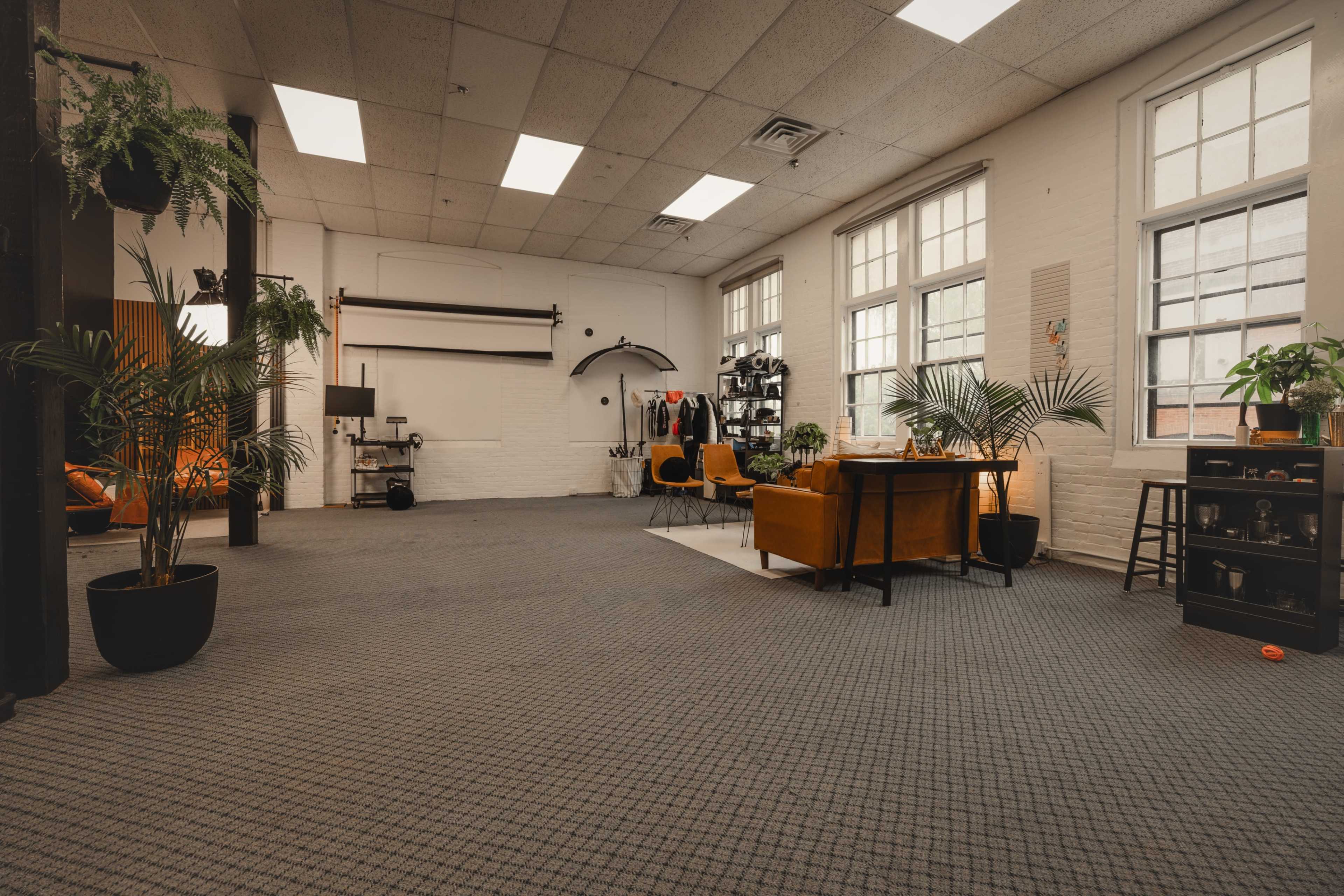 The image shows a spacious interior of a modern office or creative workspace with plants, seating areas, and large windows allowing natural light.
