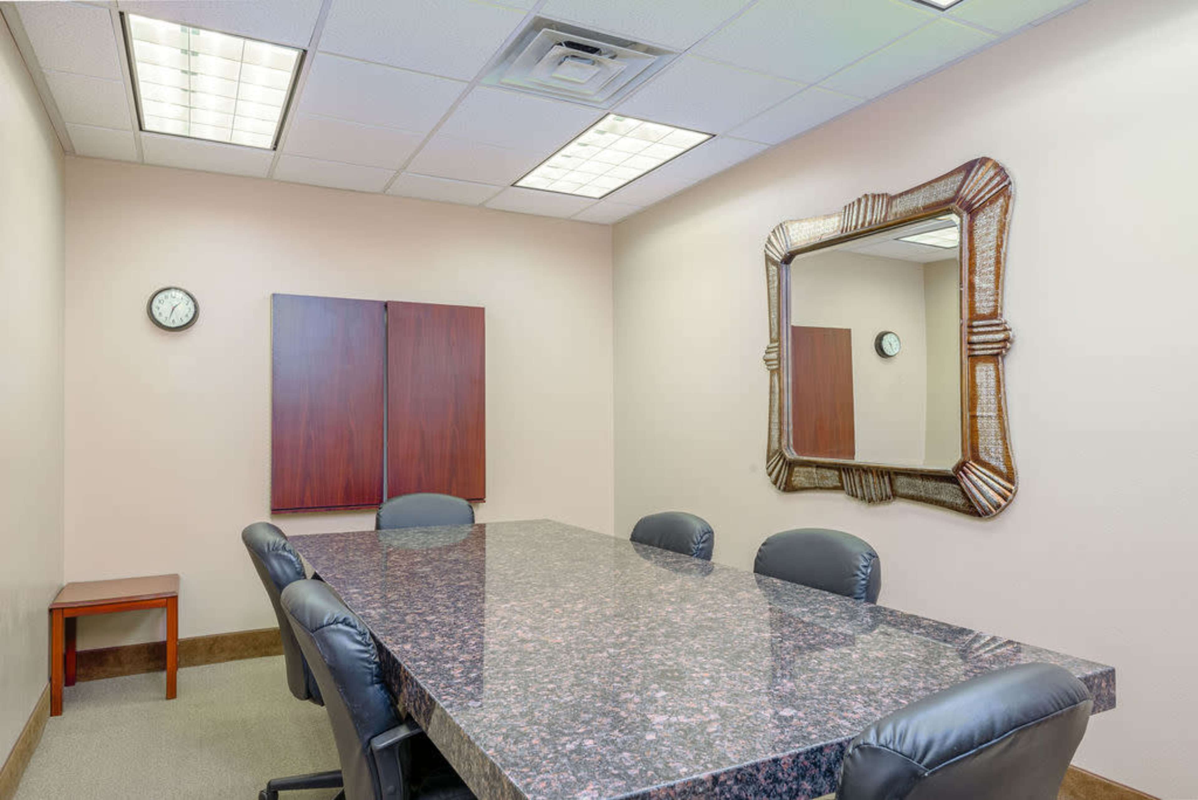 A conference room features a dark marble table surrounded by black chairs, with a clock on the wall and a large decorative mirror.