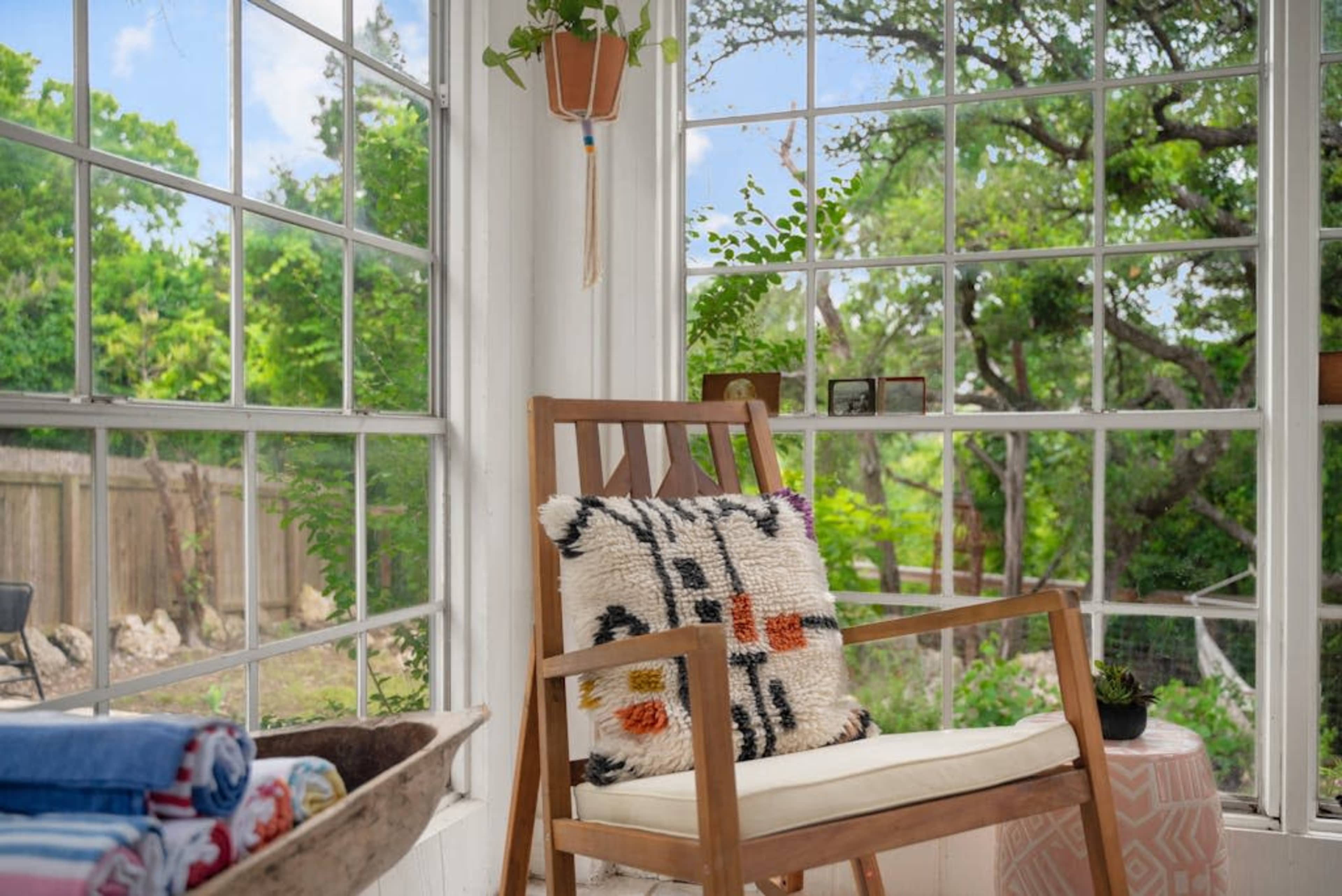 A wooden chair with a patterned cushion sits in a bright sunroom with large windows showcasing a green garden outside.