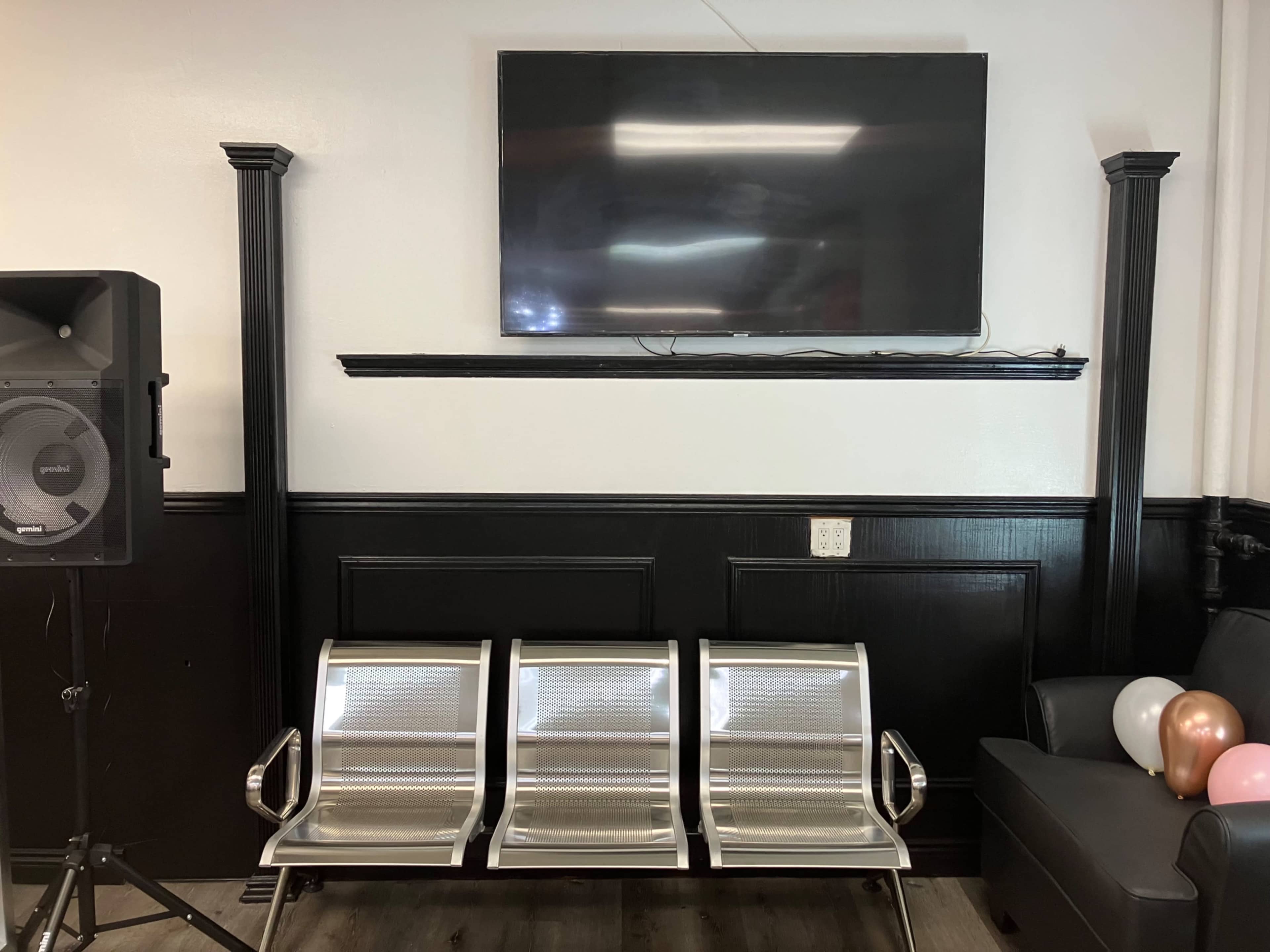 The image shows a waiting area with three metal chairs arranged in front of a wall-mounted television, alongside a speaker and a black couch with balloons.