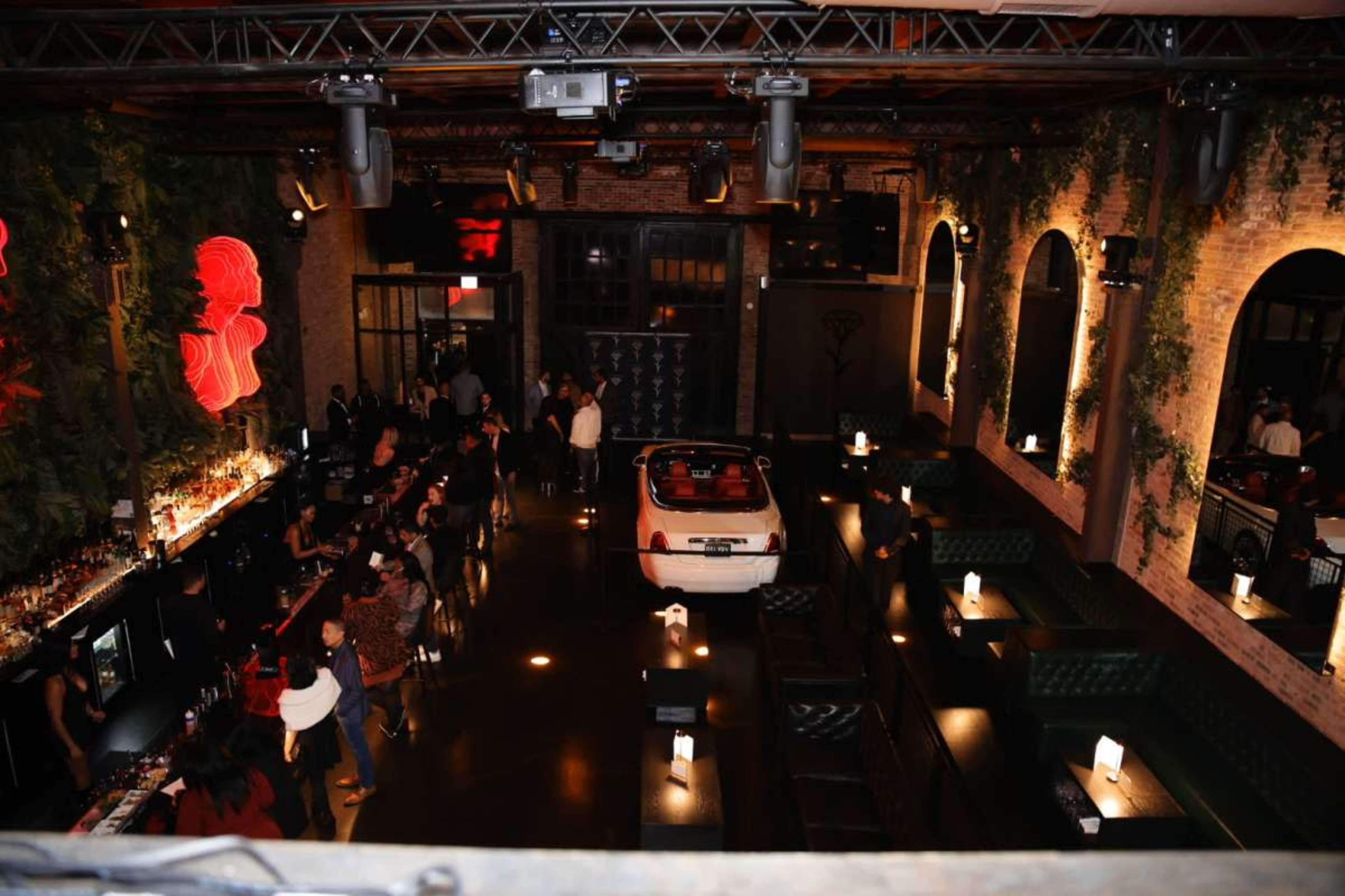 The image shows a dimly lit bar with a white car parked inside, surrounded by patrons and seating areas adorned with green upholstery and red neon lights.