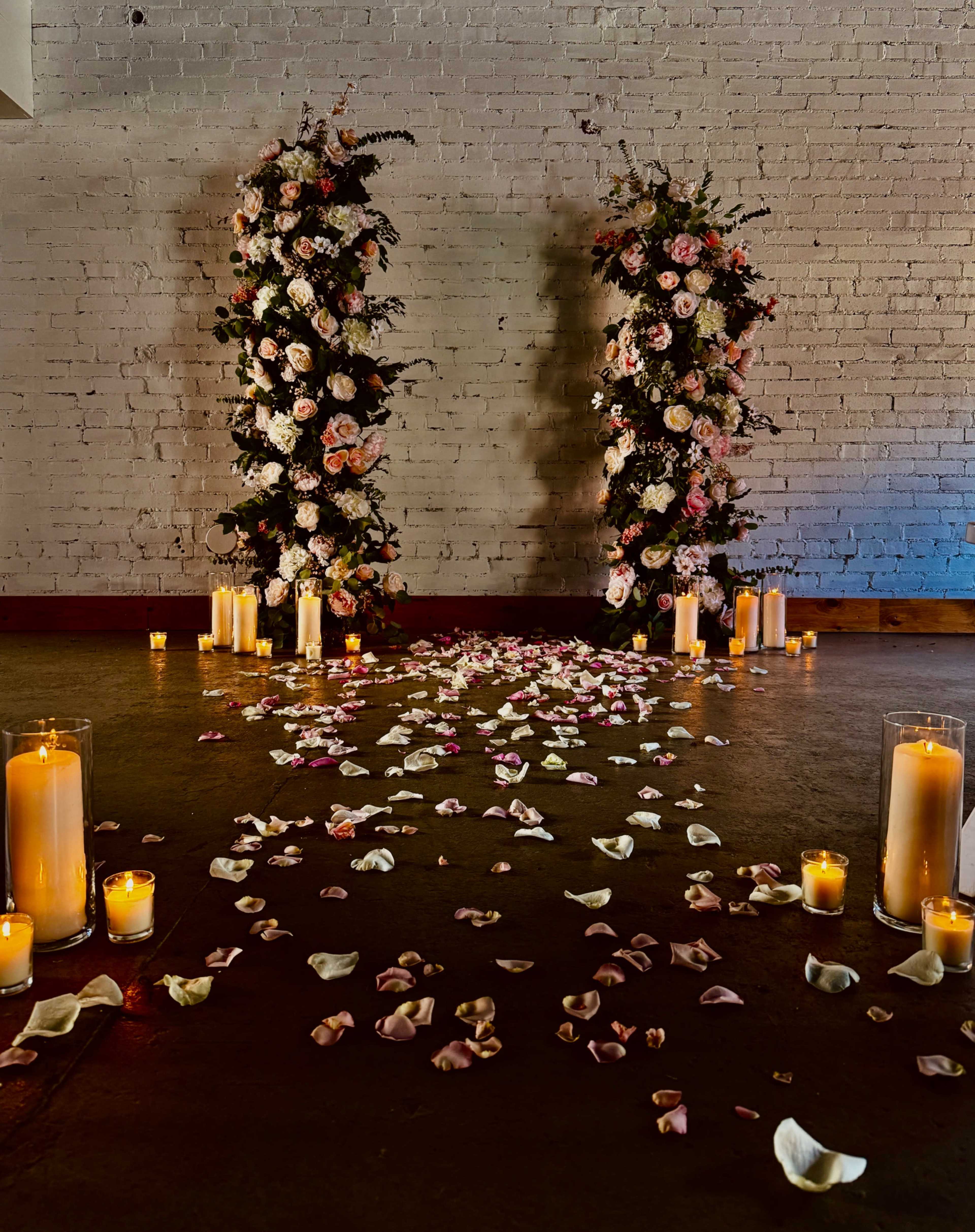 The image shows two floral arrangements on either side of a path covered with rose petals, leading to a warm glow of candles on the floor.
