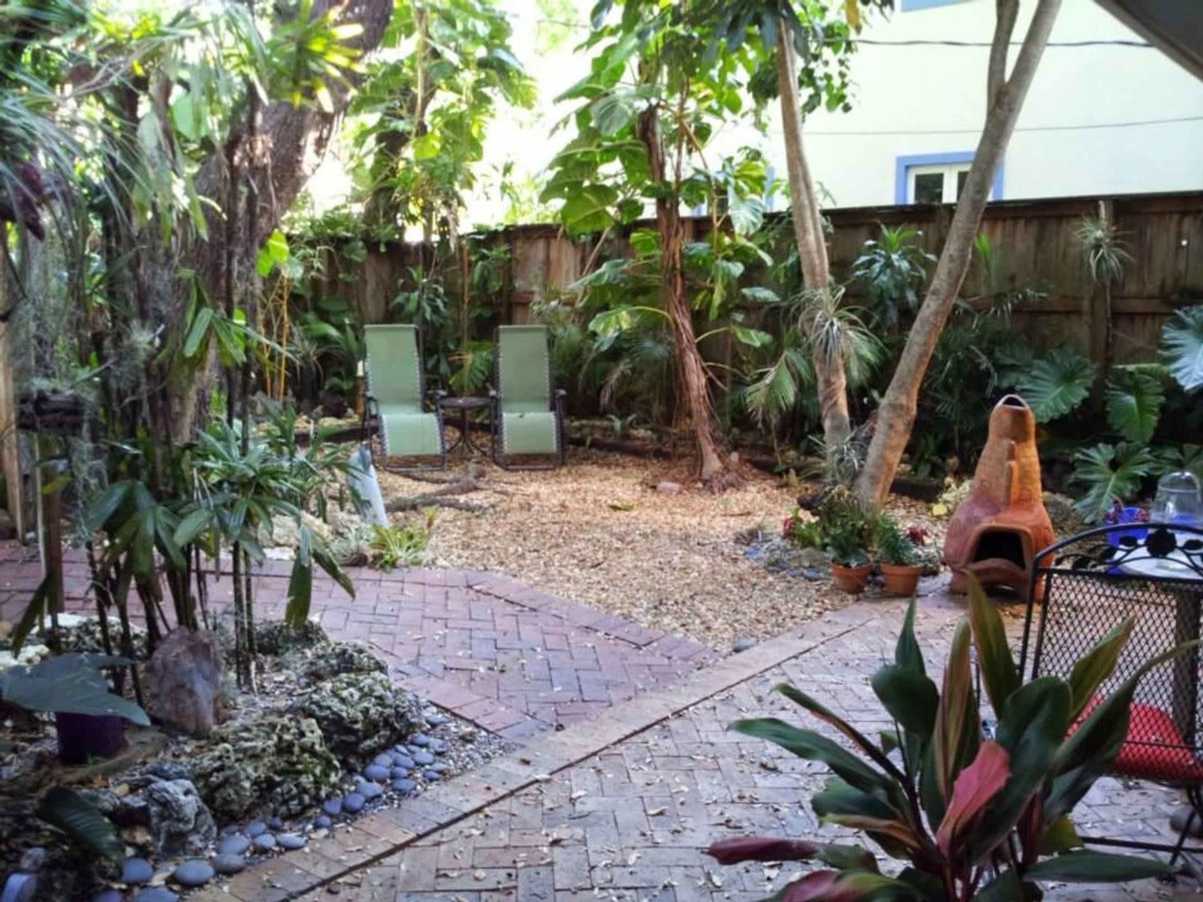 The image shows a tranquil backyard garden with stone paths, sandy areas, several potted plants, and two green lounge chairs.