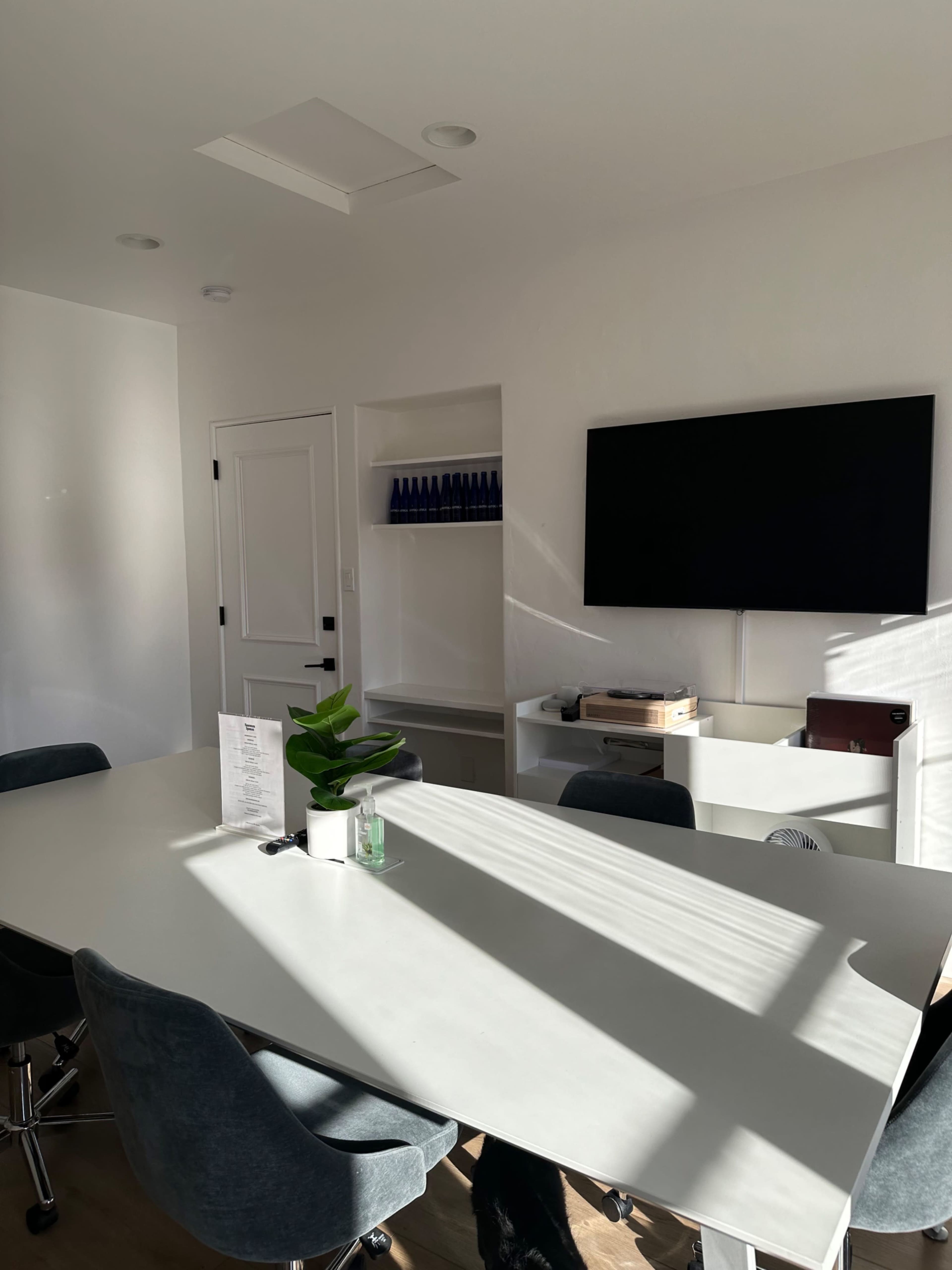 A bright, modern conference room features a large white table, chairs, a TV on the wall, and shelves with blue bottles displayed.