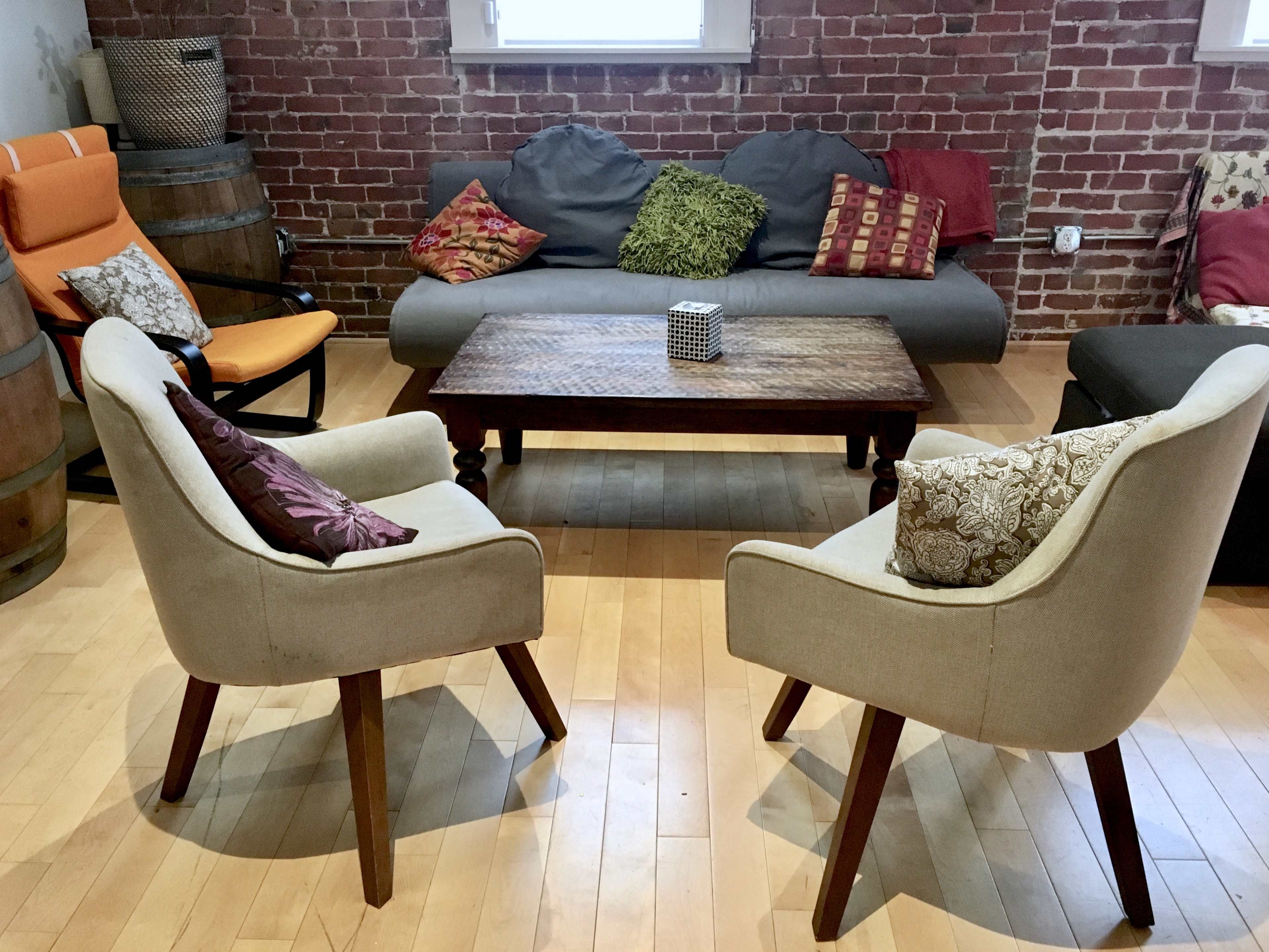 A cozy seating arrangement with two patterned chairs facing a wooden coffee table, surrounded by a sofa and colorful cushions against a brick wall backdrop.