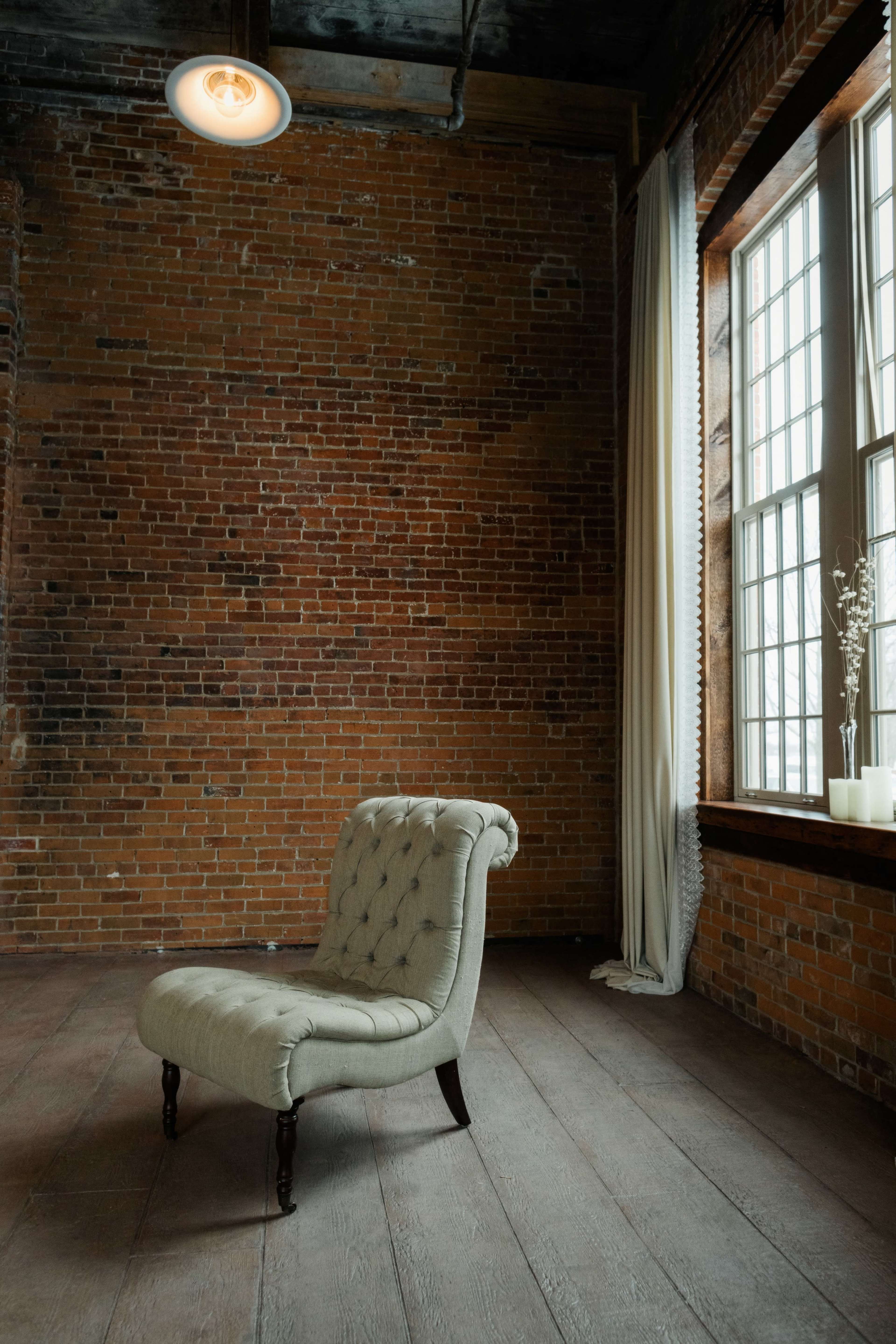 A tufted upholstered chair is positioned in a room with exposed brick walls and large windows.