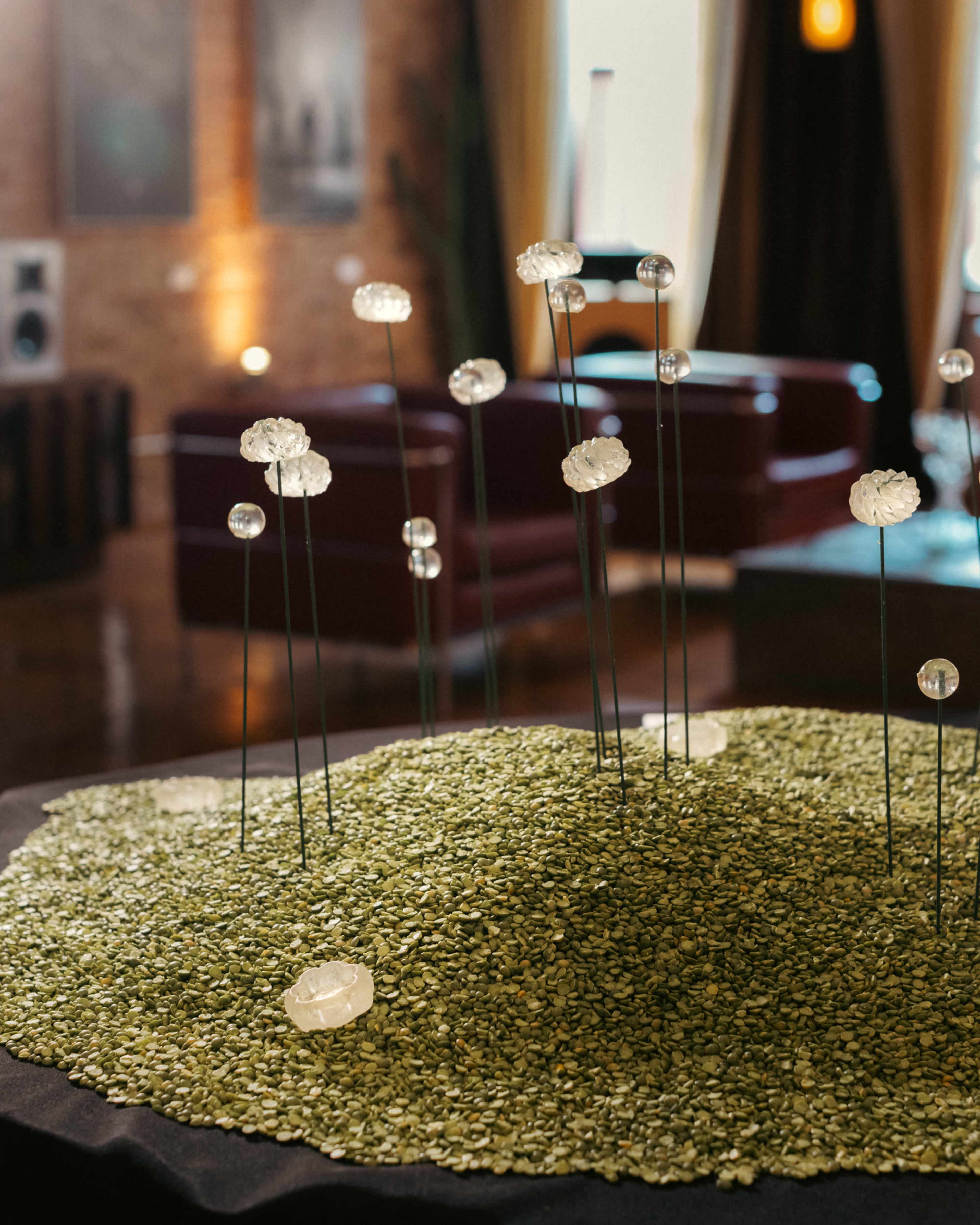 A decorative arrangement featuring glass flowers atop a bed of green stones is displayed in a stylish interior space with couches and artwork in the background.