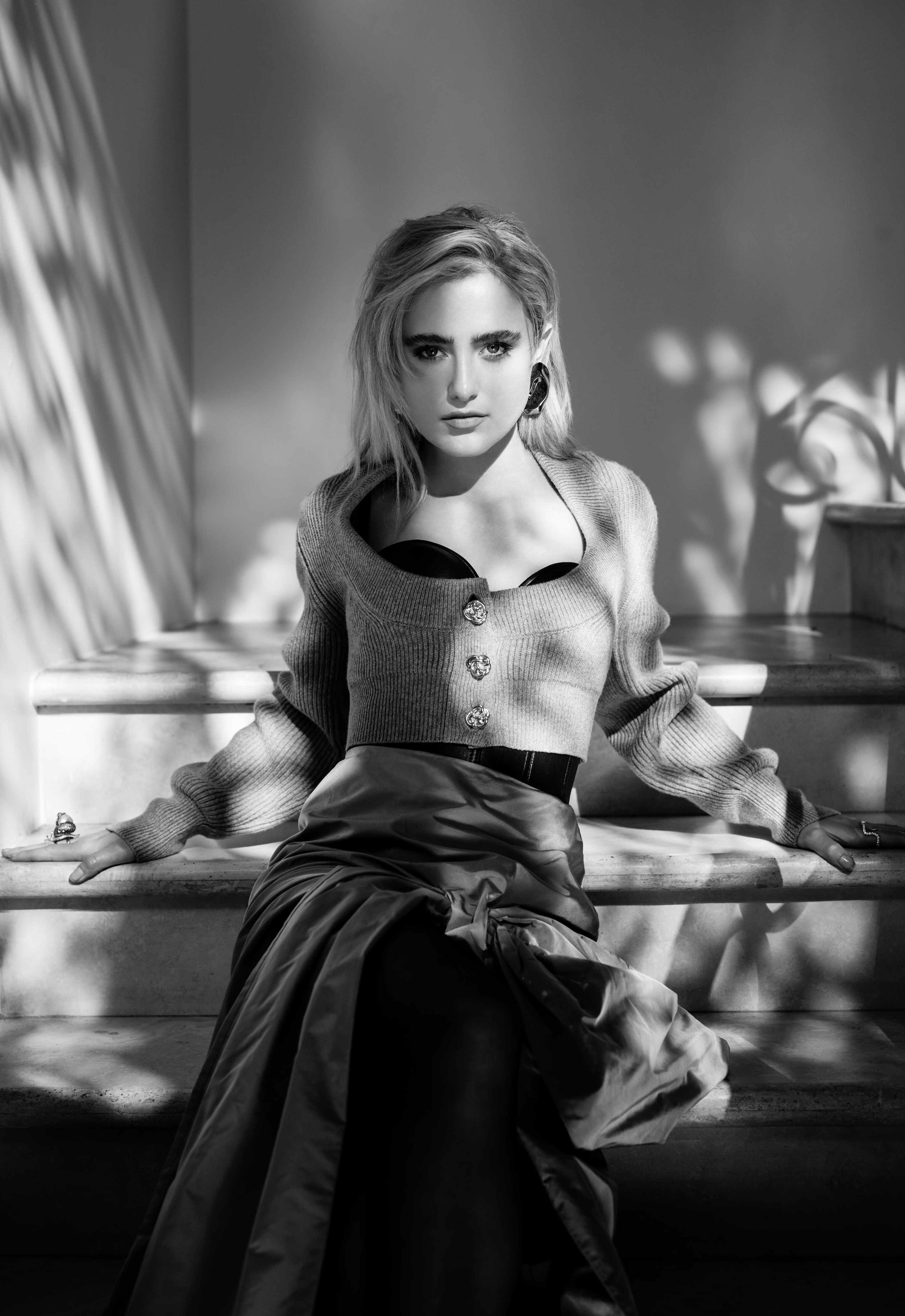 A person sits on a staircase, wearing a fitted top and a long skirt, with light and shadow creating patterns on the wall behind them.