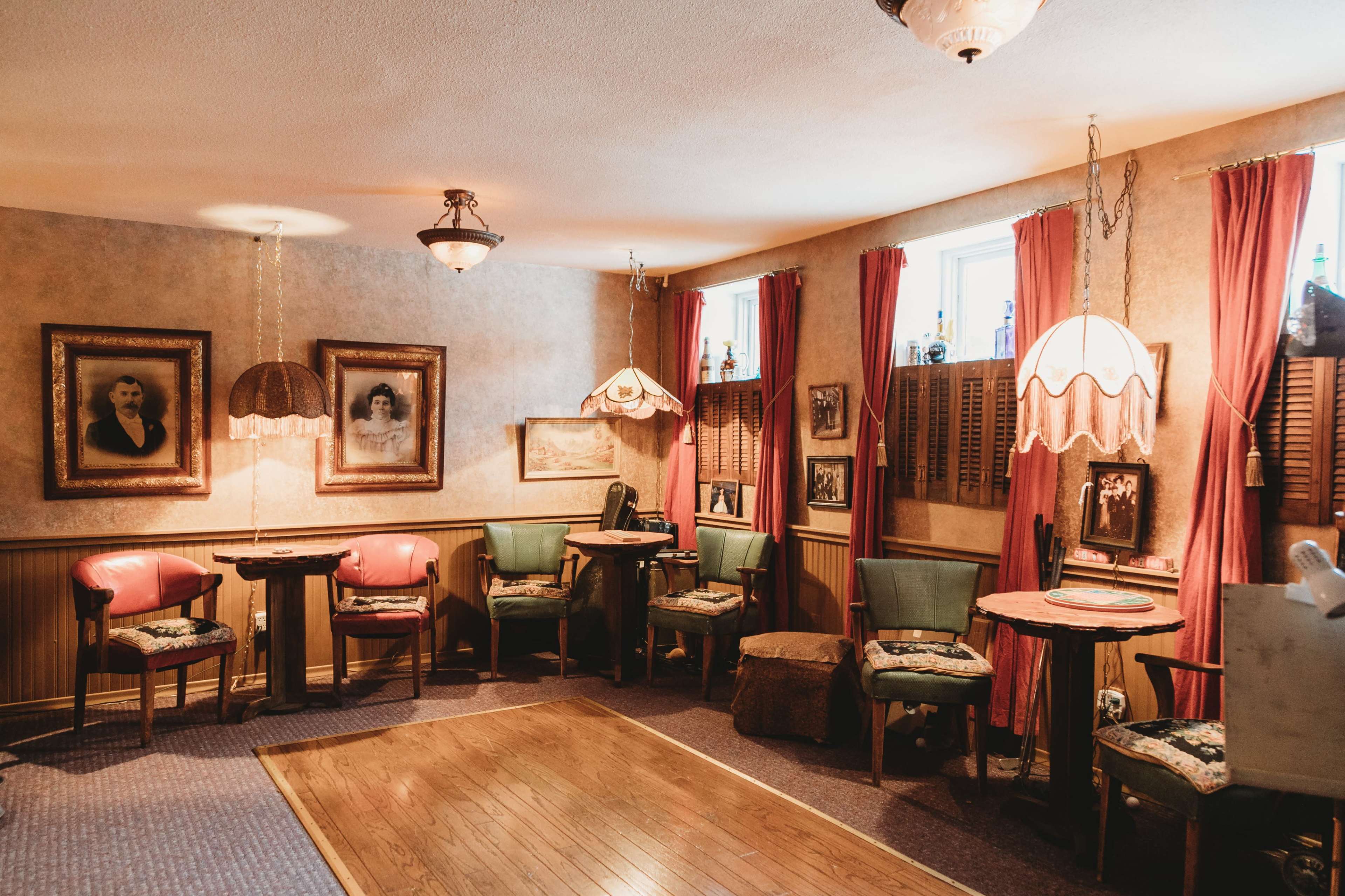 The image shows a vintage room with framed pictures on the walls, wooden tables, and upholstered chairs arranged around a wooden floor.