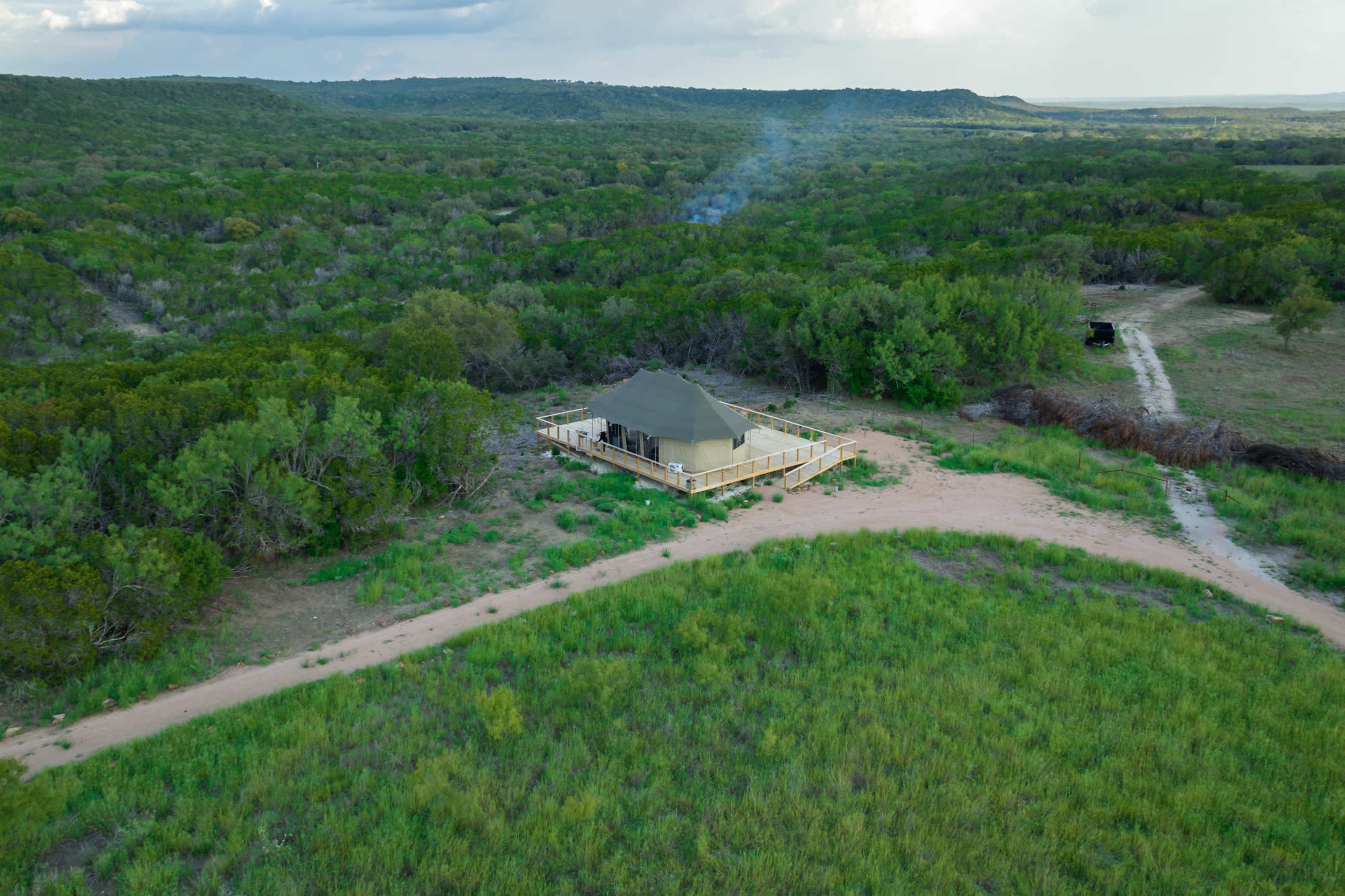 A tented lodge is situated on a dirt path surrounded by lush greenery and open fields.