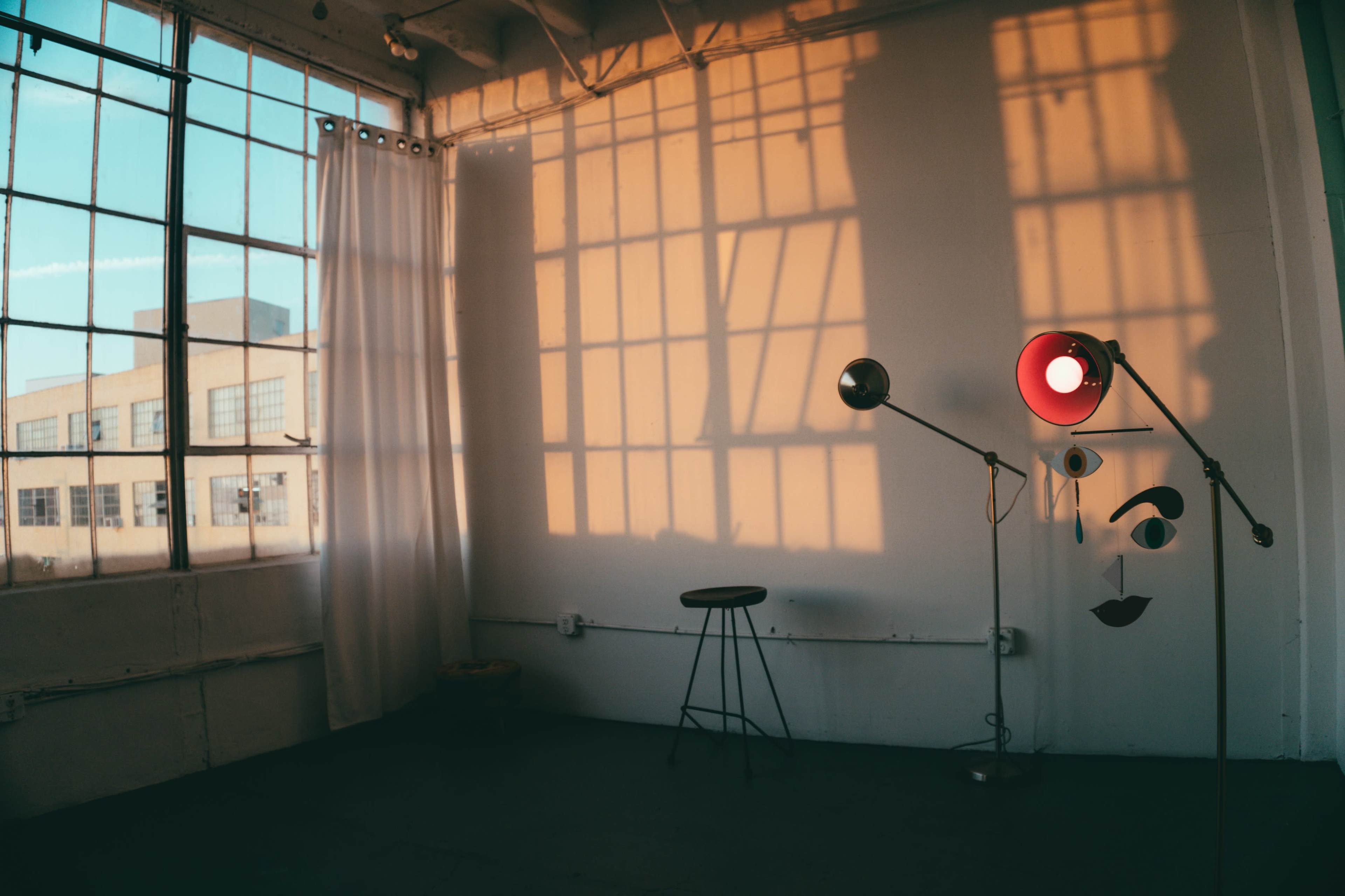 The image shows a minimalistic room with large windows, a stool, and a floor lamp casting shadows on a white wall.