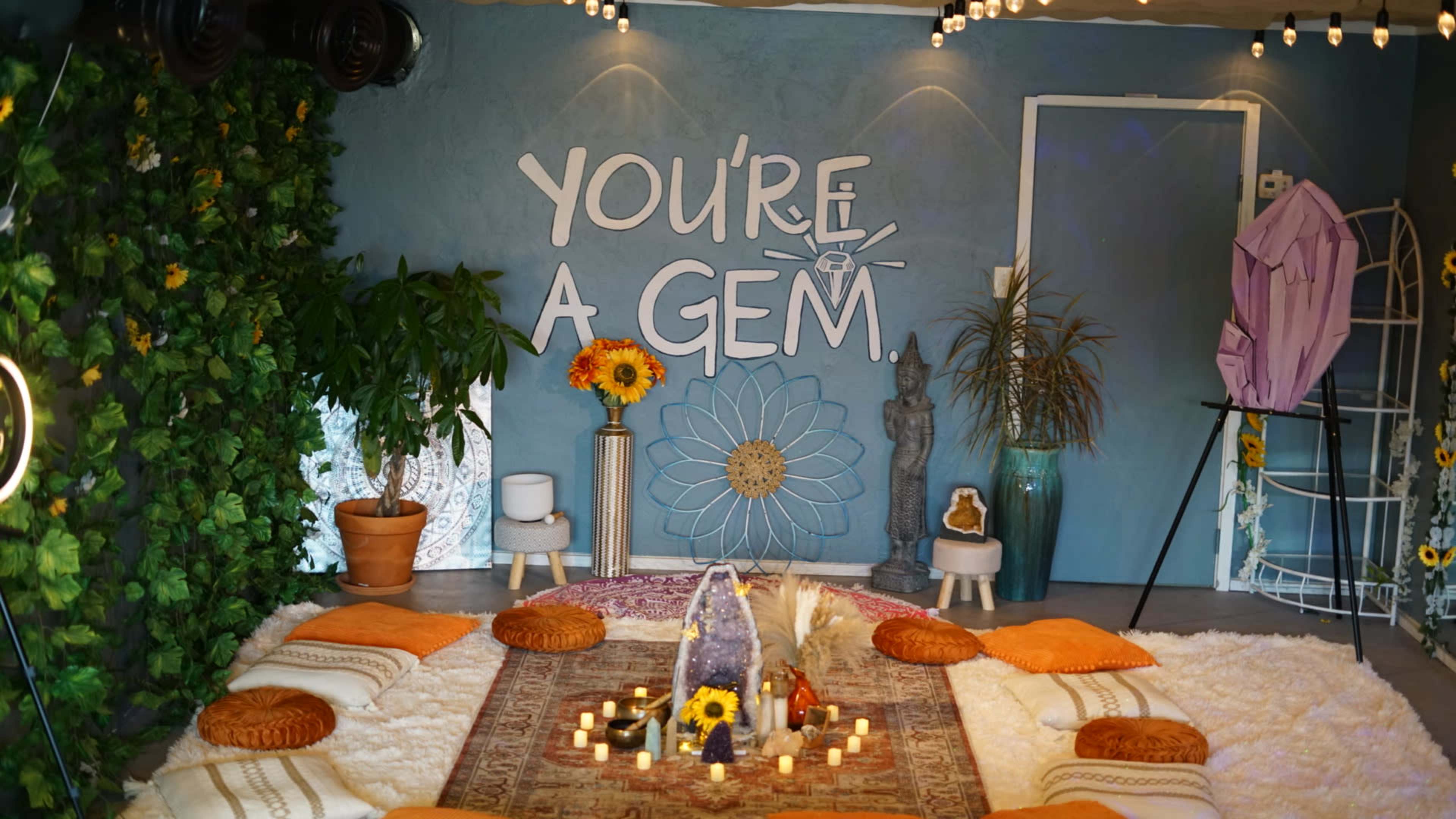 A cozy interior space features a central rug surrounded by cushions, with a decorative wall that reads "YOU'RE A GEM" and various plants and candles throughout.