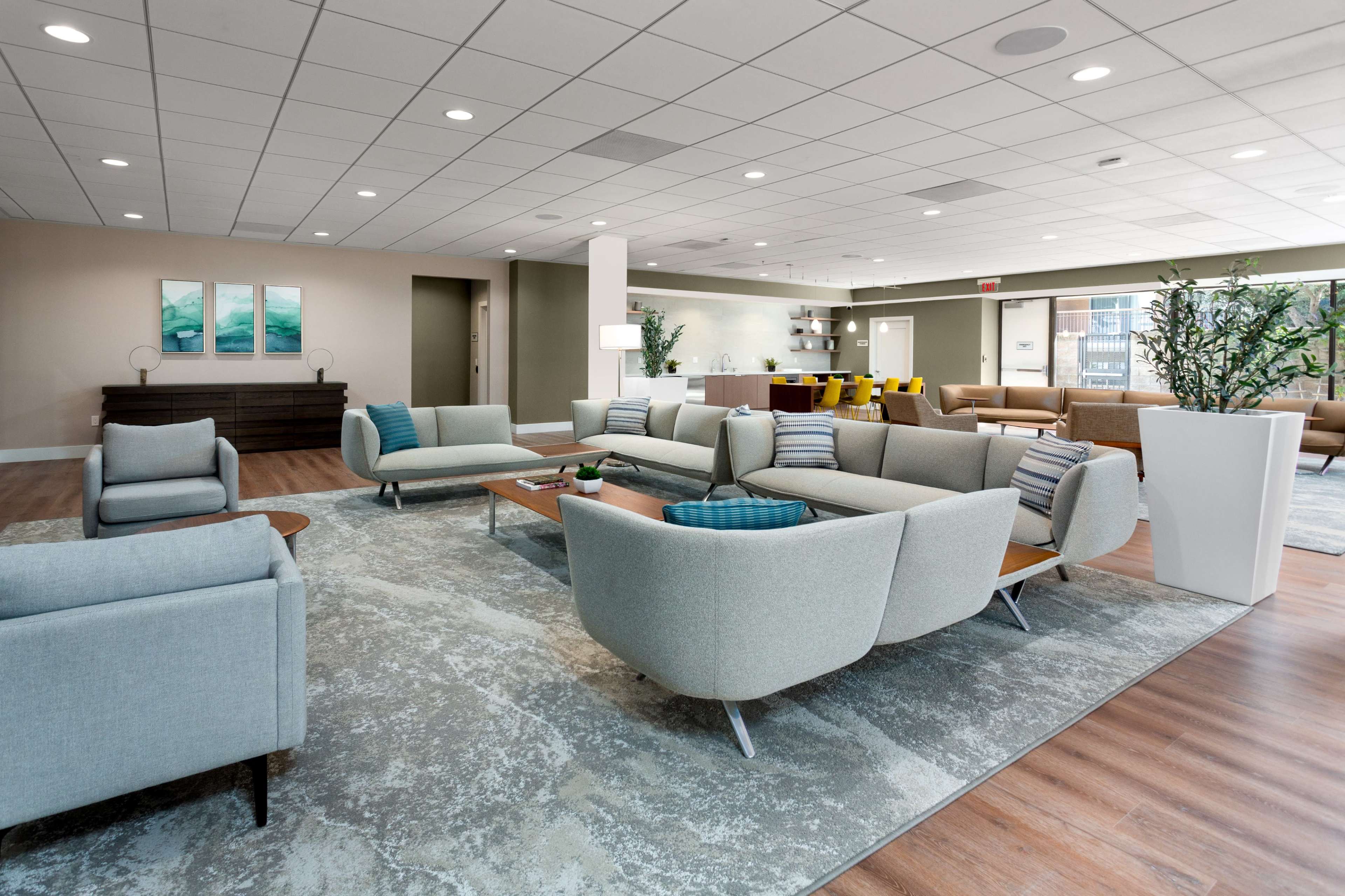 A spacious, modern lounge area with light-colored furniture arranged around a central table on a patterned rug.