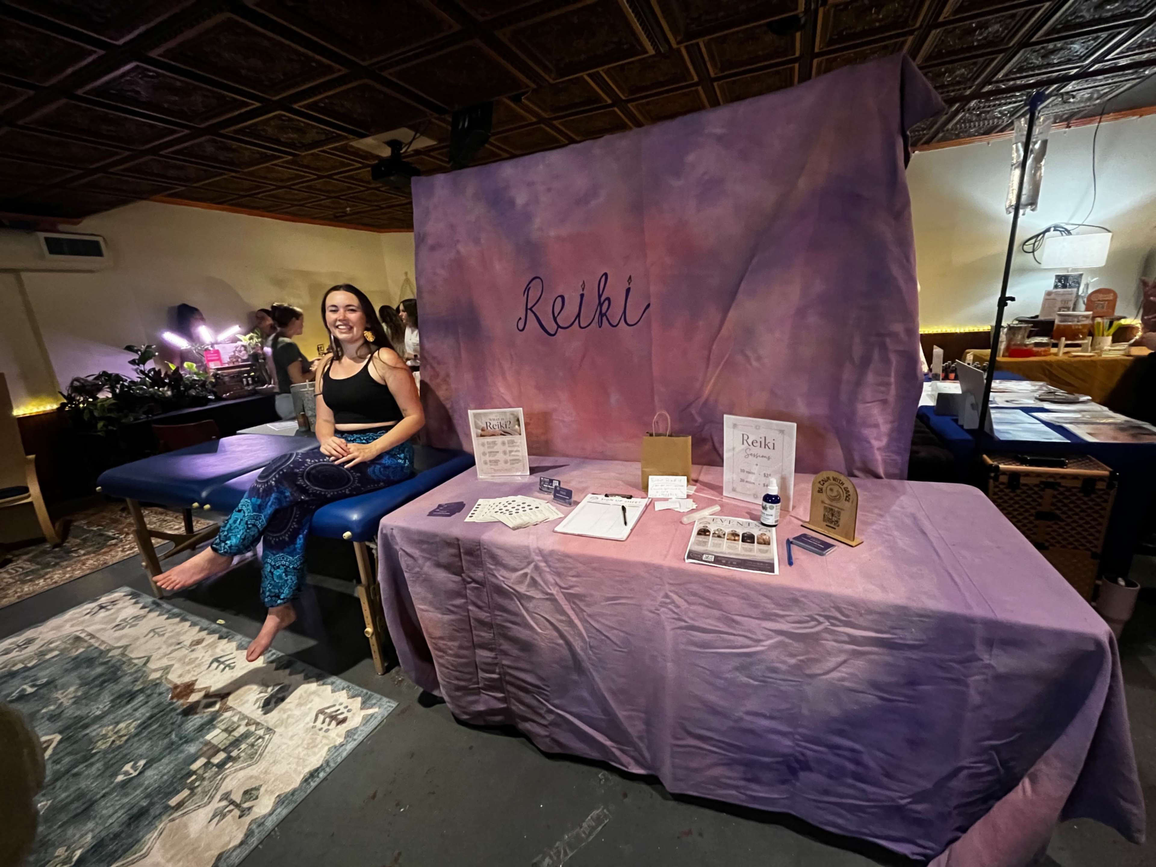A woman sits at a table covered with a purple cloth, displaying materials related to Reiki, in a softly lit room.