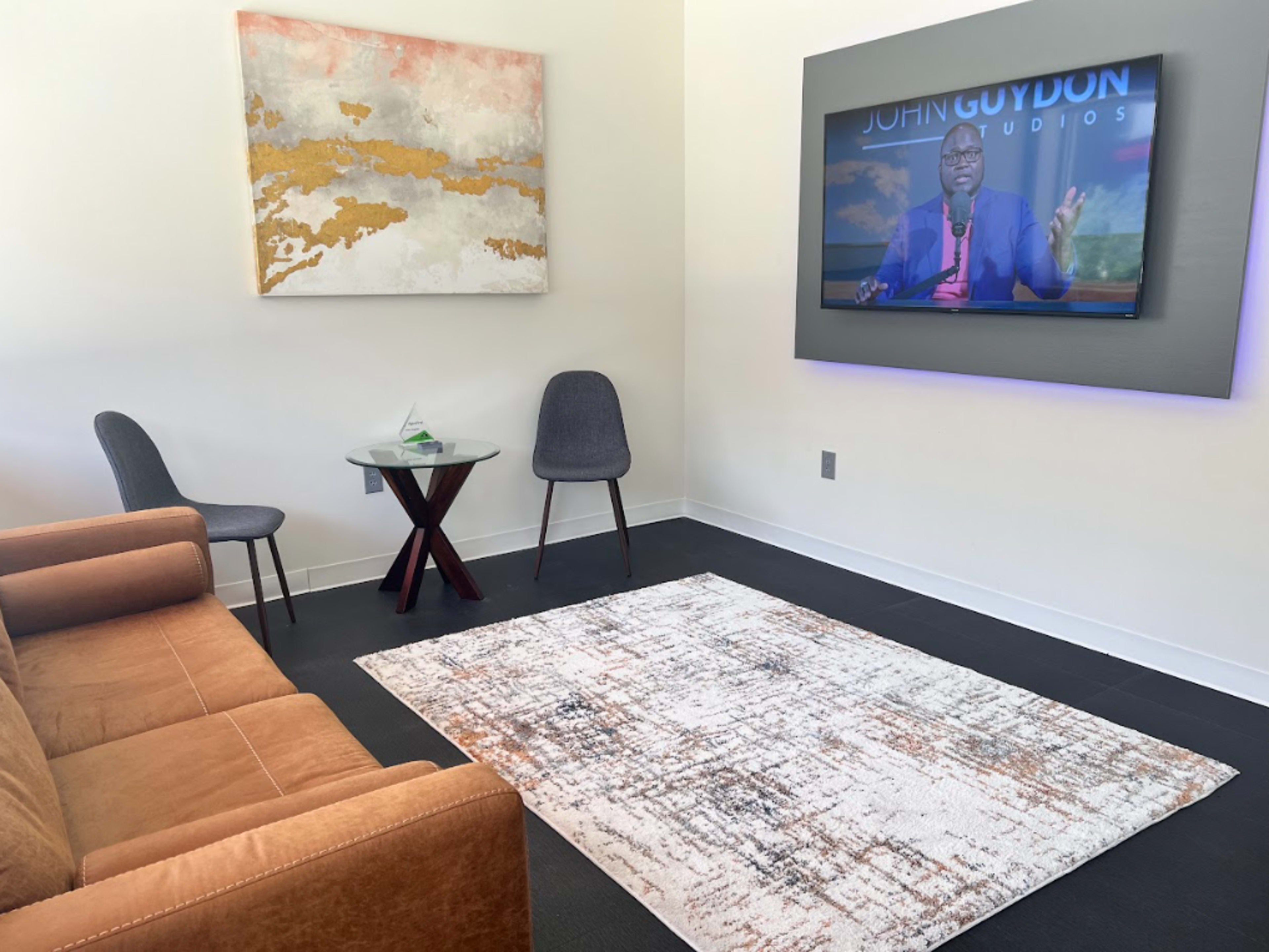 The image shows a modern sitting area with a brown sofa, a round wooden table, two chairs, a patterned rug, a wall-mounted television displaying a program, and artwork on the wall.